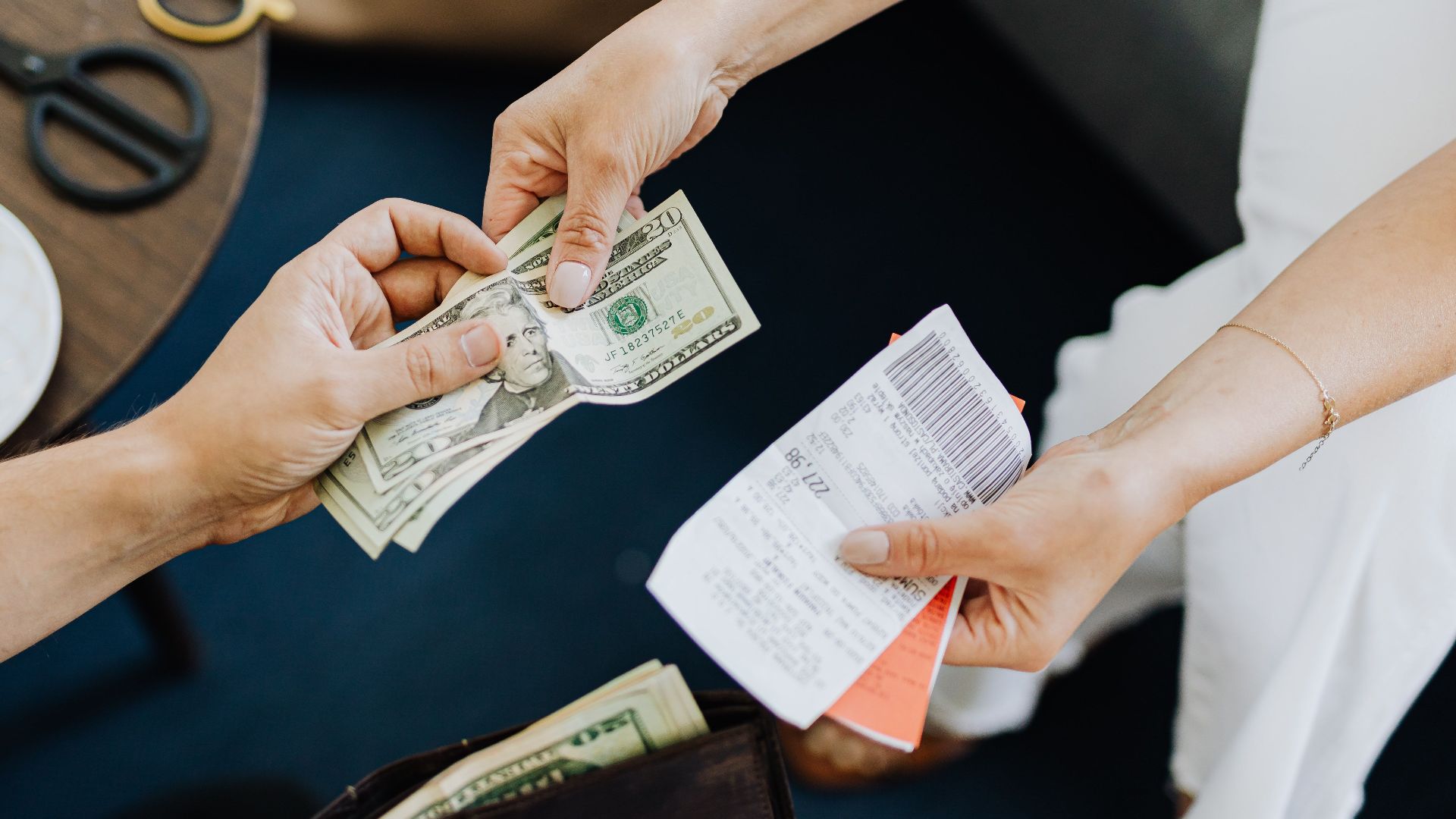 Close-up of a transaction with US dollar bills and receipts being exchanged over a table with coffee and scissors.