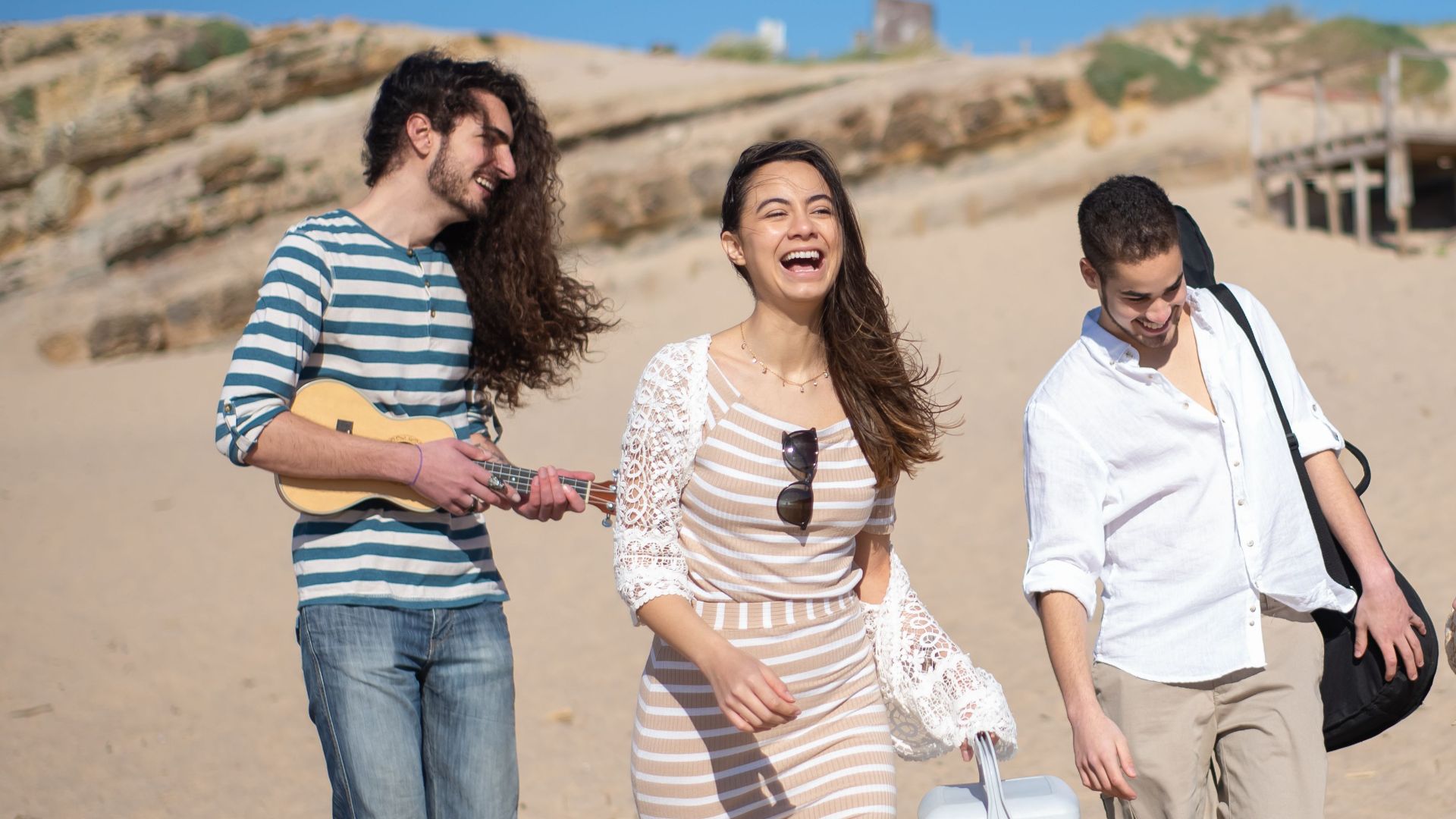 A group of friends laughing and walking barefoot on a sunny beach day in Portugal.
