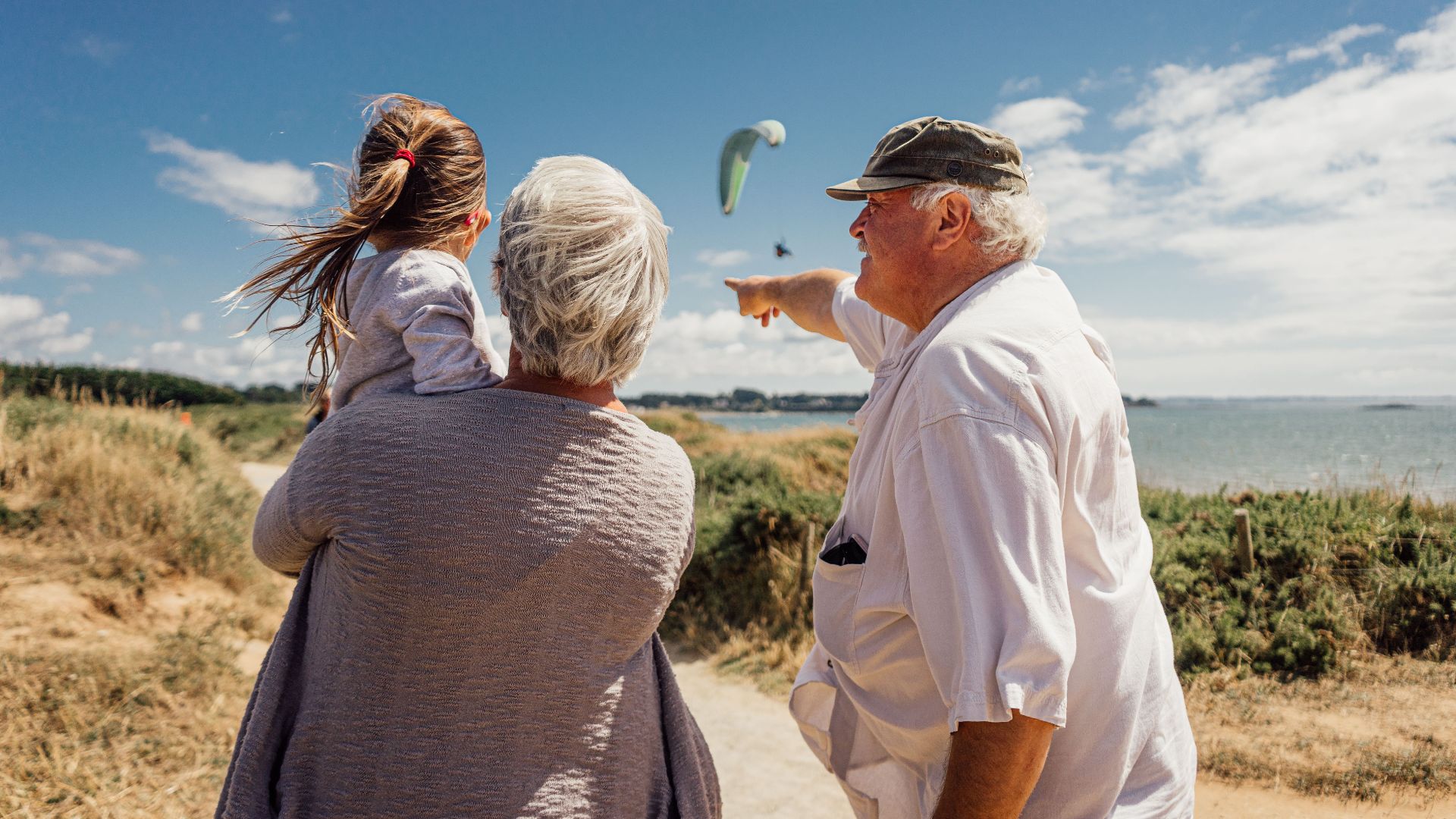 Elderly couple with granddaughter watching the sea and a paraglider on a sunny day in Pénestin, Brittany, France.