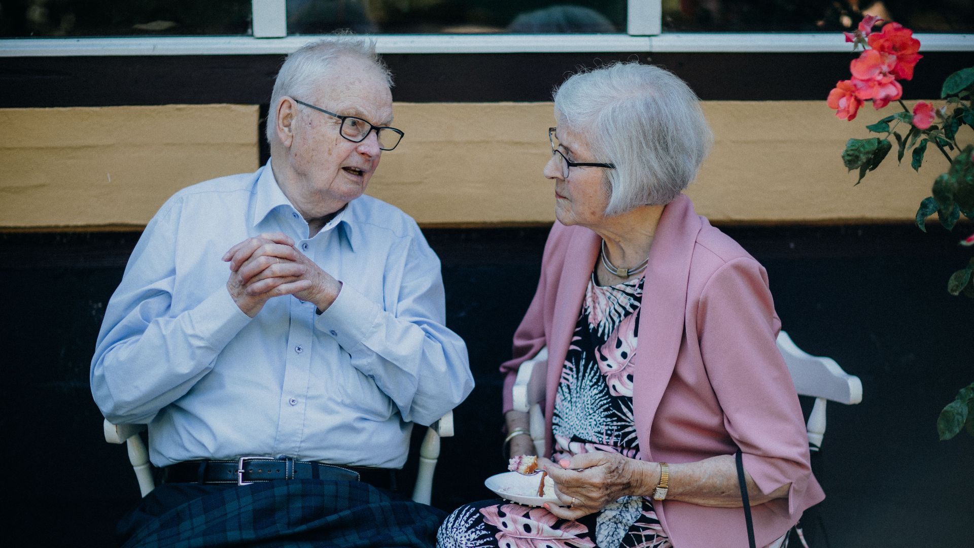 Senior couple sitting outside, engaged in conversation with flowers nearby. Captures a candid moment.