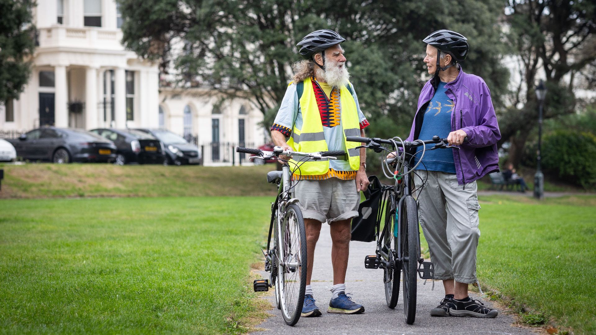 Two senior men with bicycles and helmets, enjoying a talk in a scenic outdoor park.