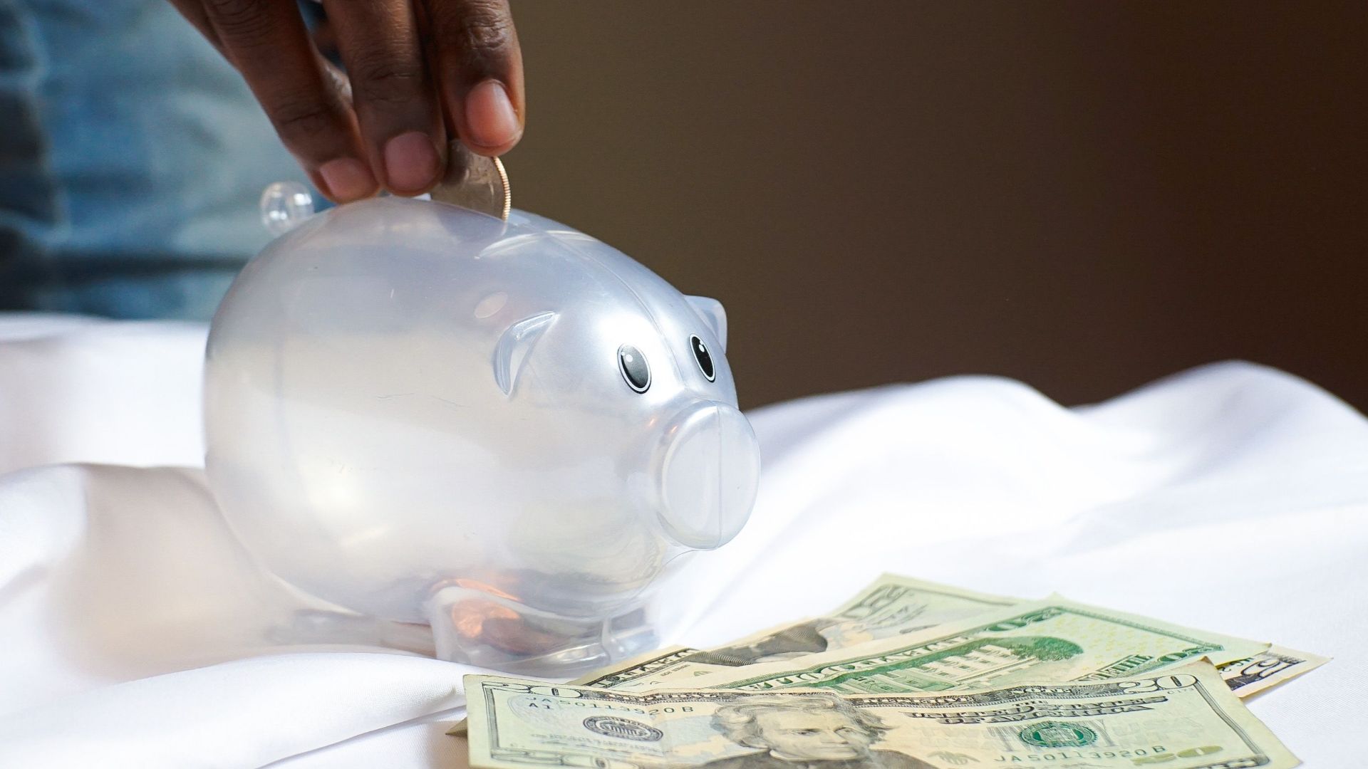 A hand placing a coin in a piggy bank with dollar bills nearby signifying savings and finance.