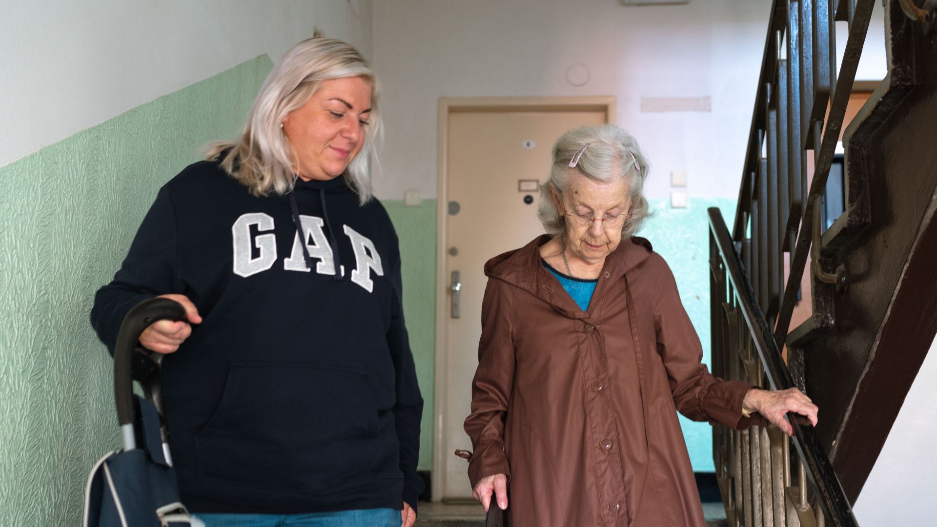 A senior woman assisted by a caregiver walking down indoor stairs in Prague.