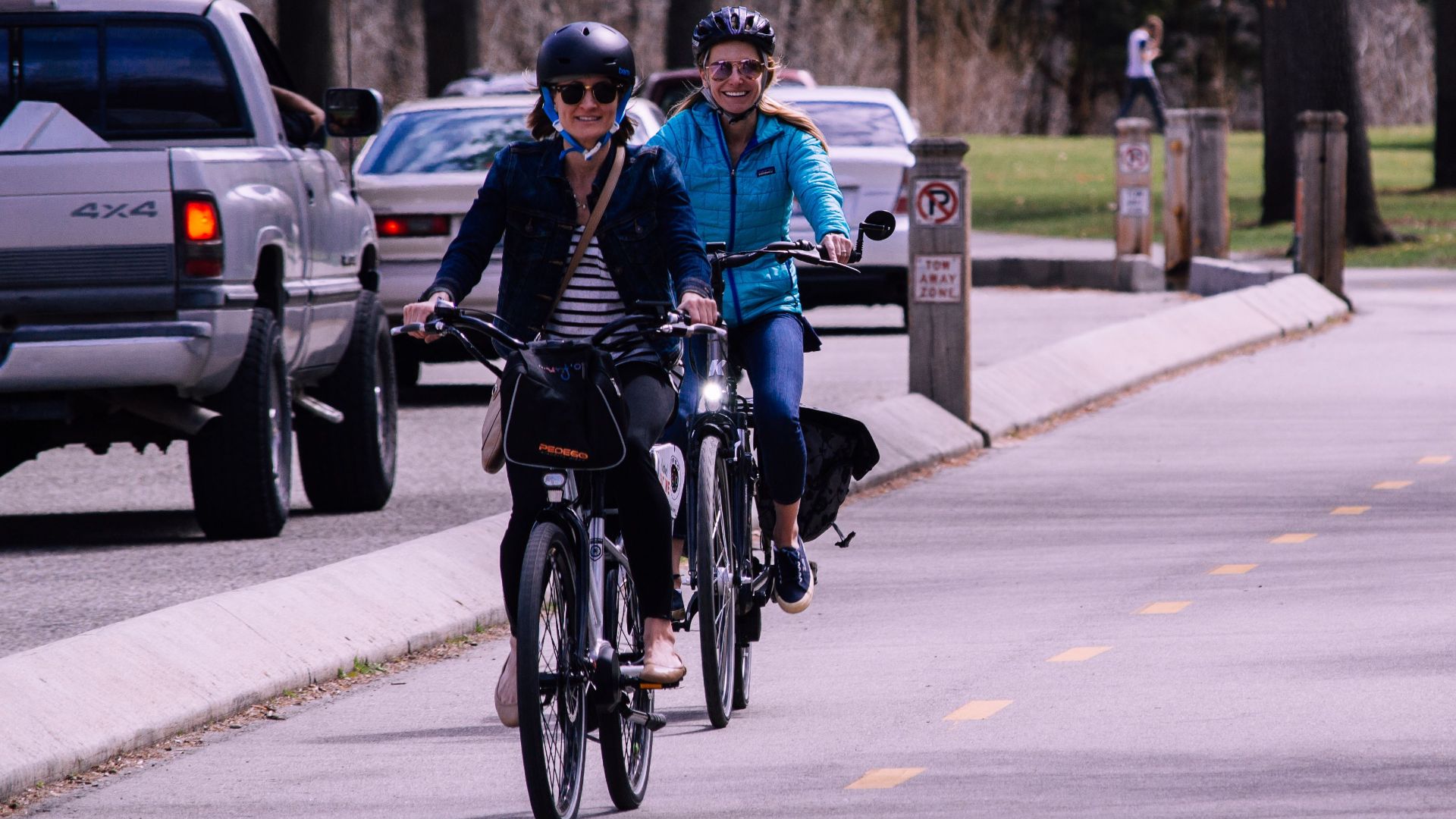Two women cycling on an urban bike path in a park with vehicles nearby.