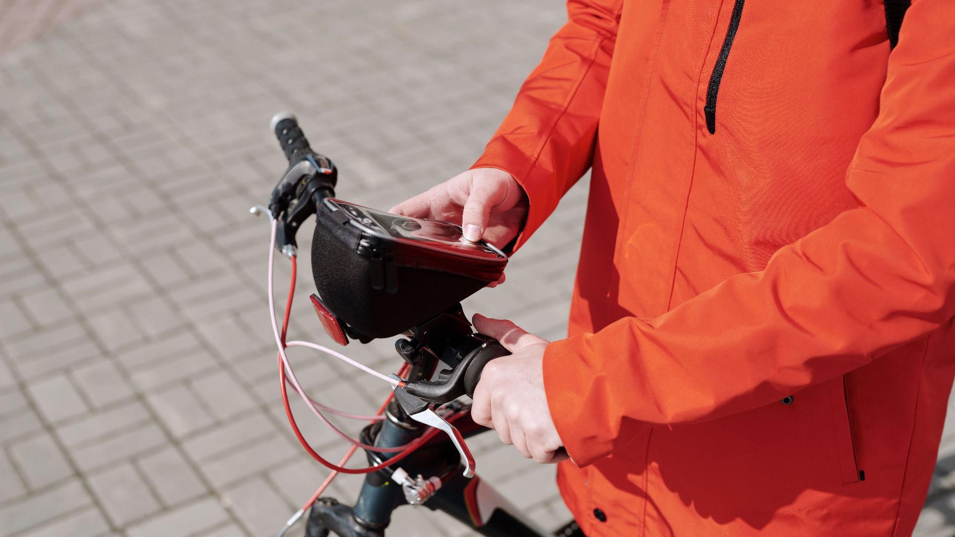 A food delivery cyclist in an orange jacket uses a smartphone while riding in the city.