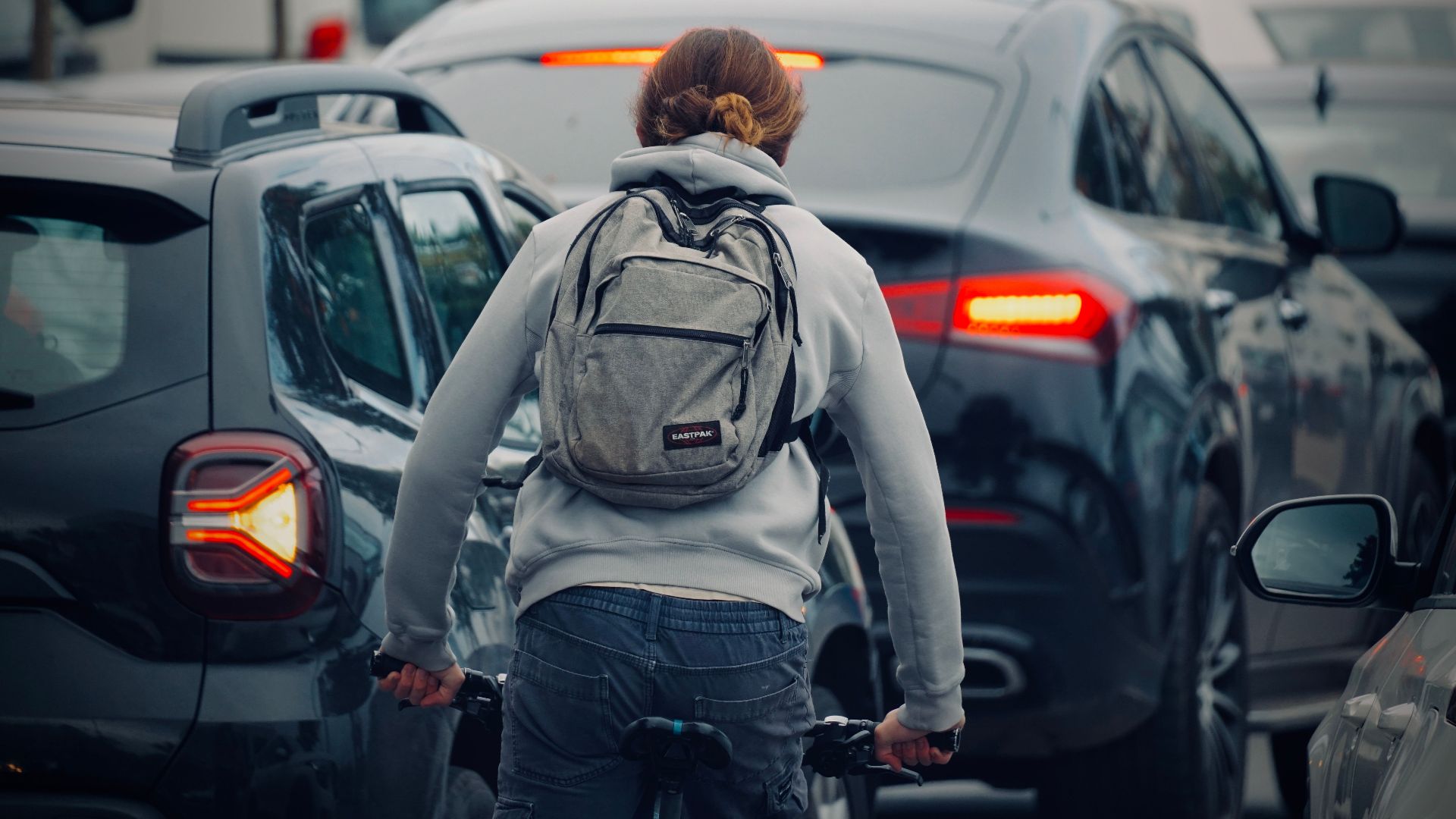 A cyclist wearing a backpack rides through a congested city traffic jam during daytime.