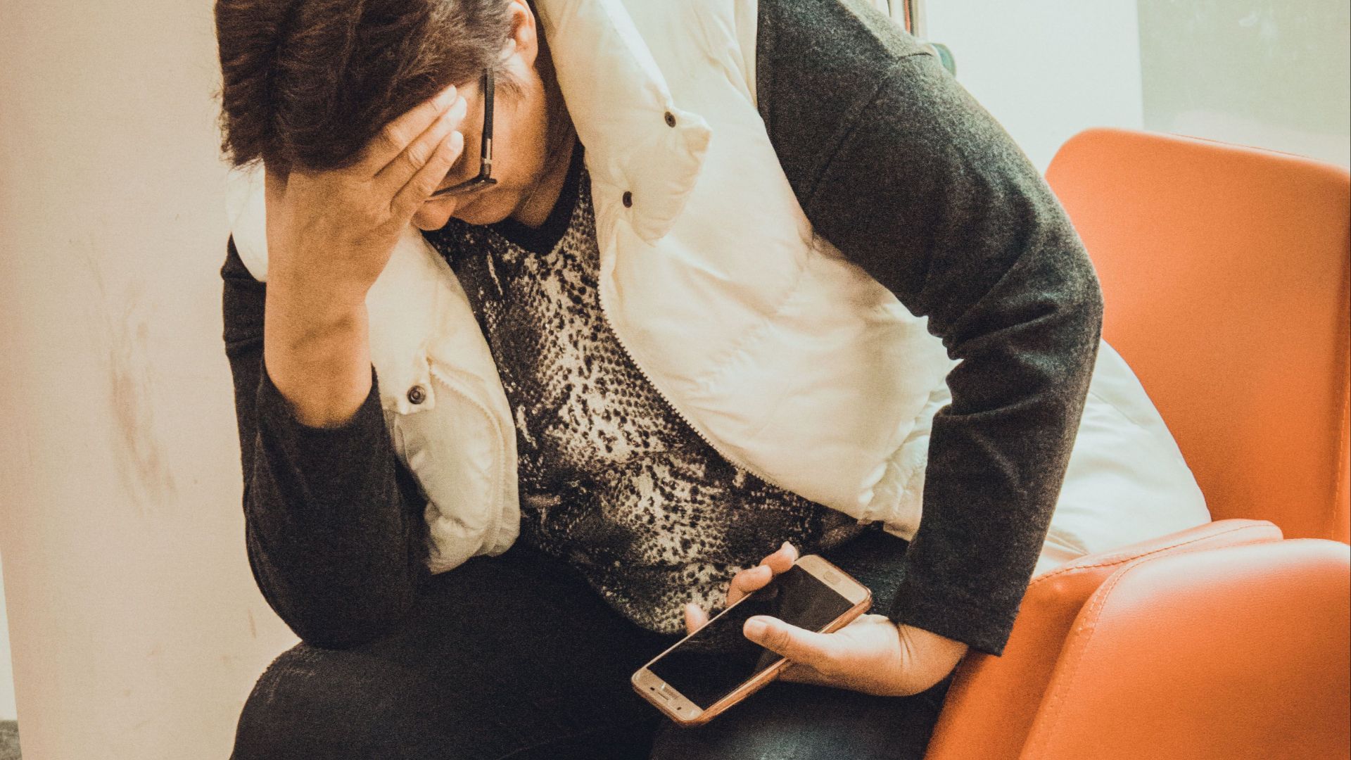 An anxious woman sits in a waiting room, holding her smartphone and resting her head on her hand.