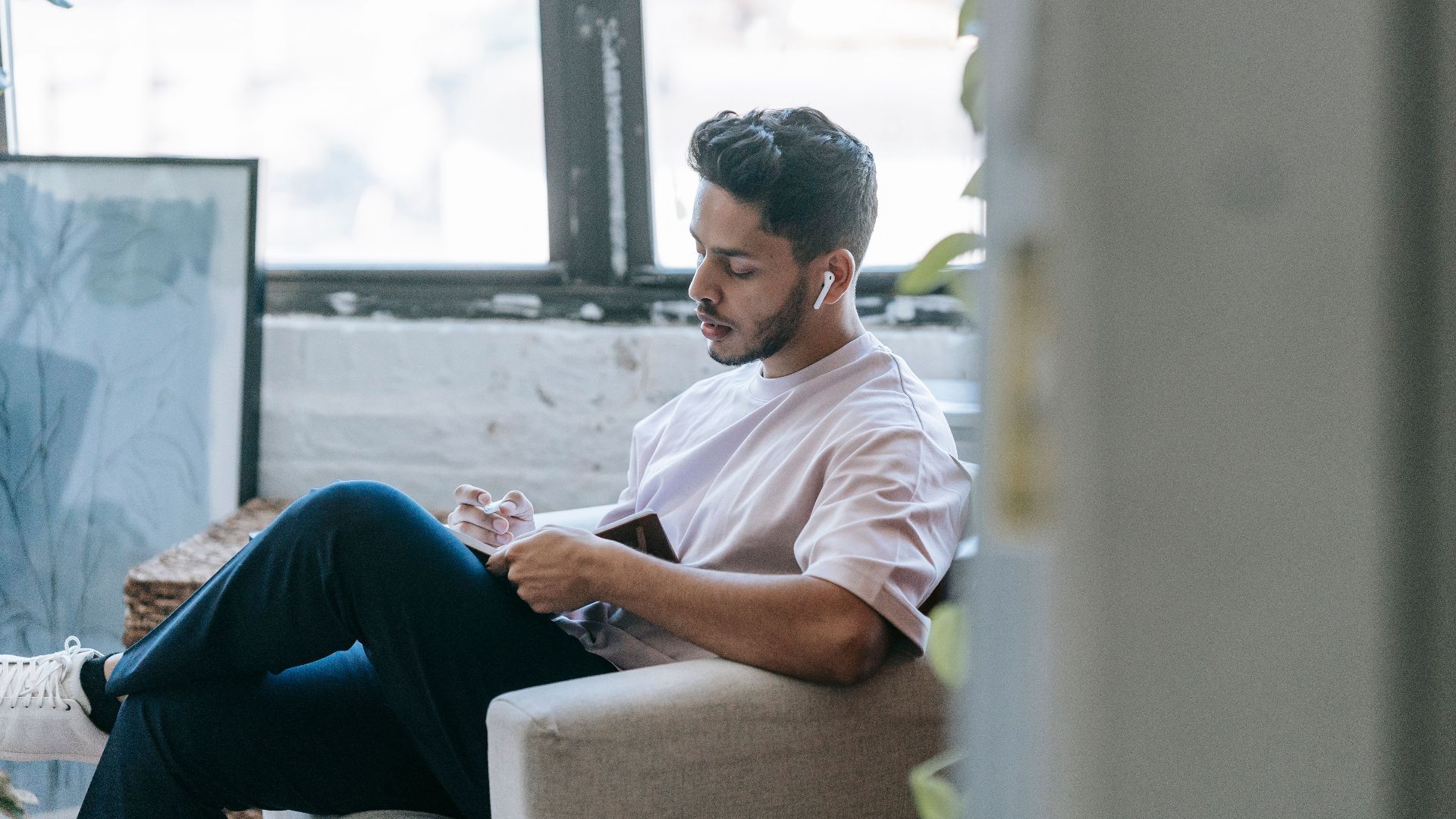 Pensive young ethnic male in casual wear writing in diary and listening to music via earbuds while sitting in cozy armchair