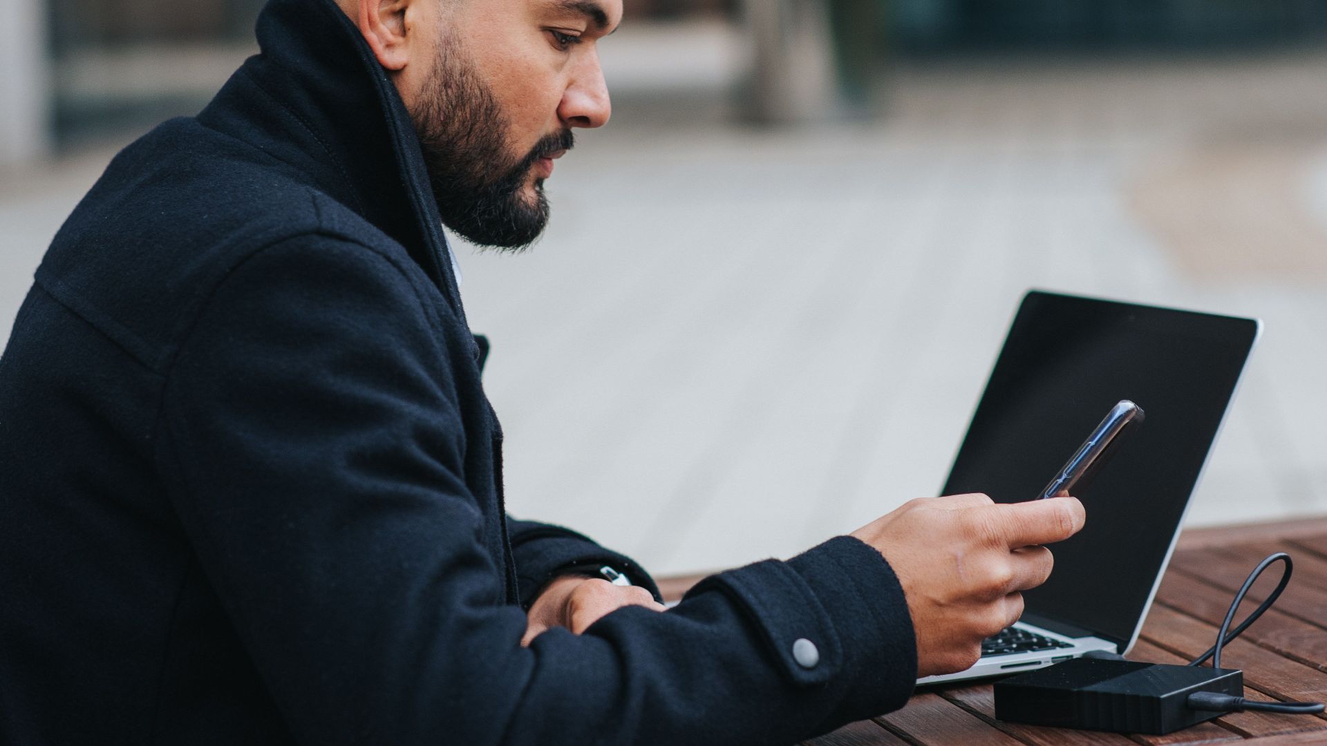 Side view of young concentrated ethnic male manager surfing internet on cellphone at cafe table with netbook and power bank on street