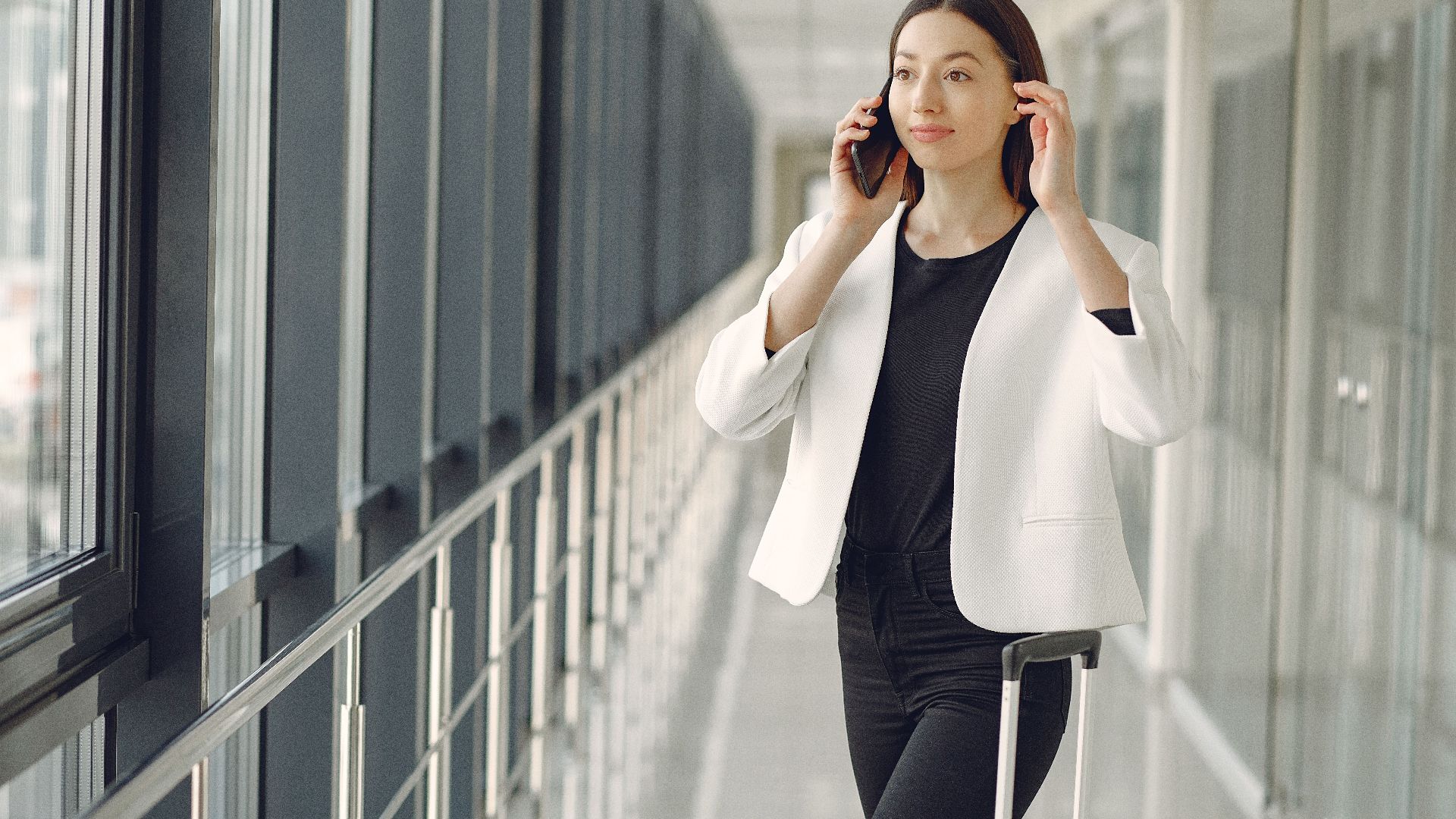 Positive young businesswoman in stylish formal wear with suitcase talking on smartphone and standing in modern airport corridor while waiting for flight