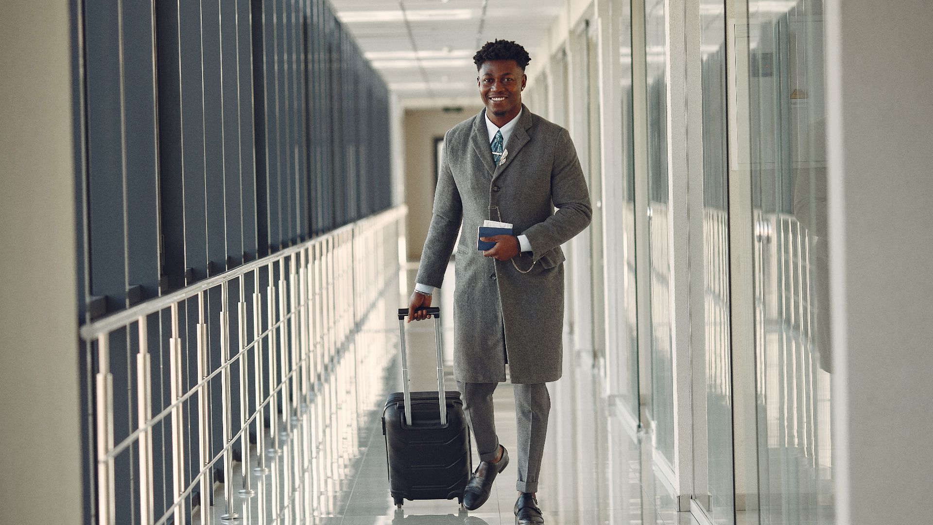 Full length of happy African American male in stylish classy gray clothes with suitcase and passport walking in contemporary airport terminal