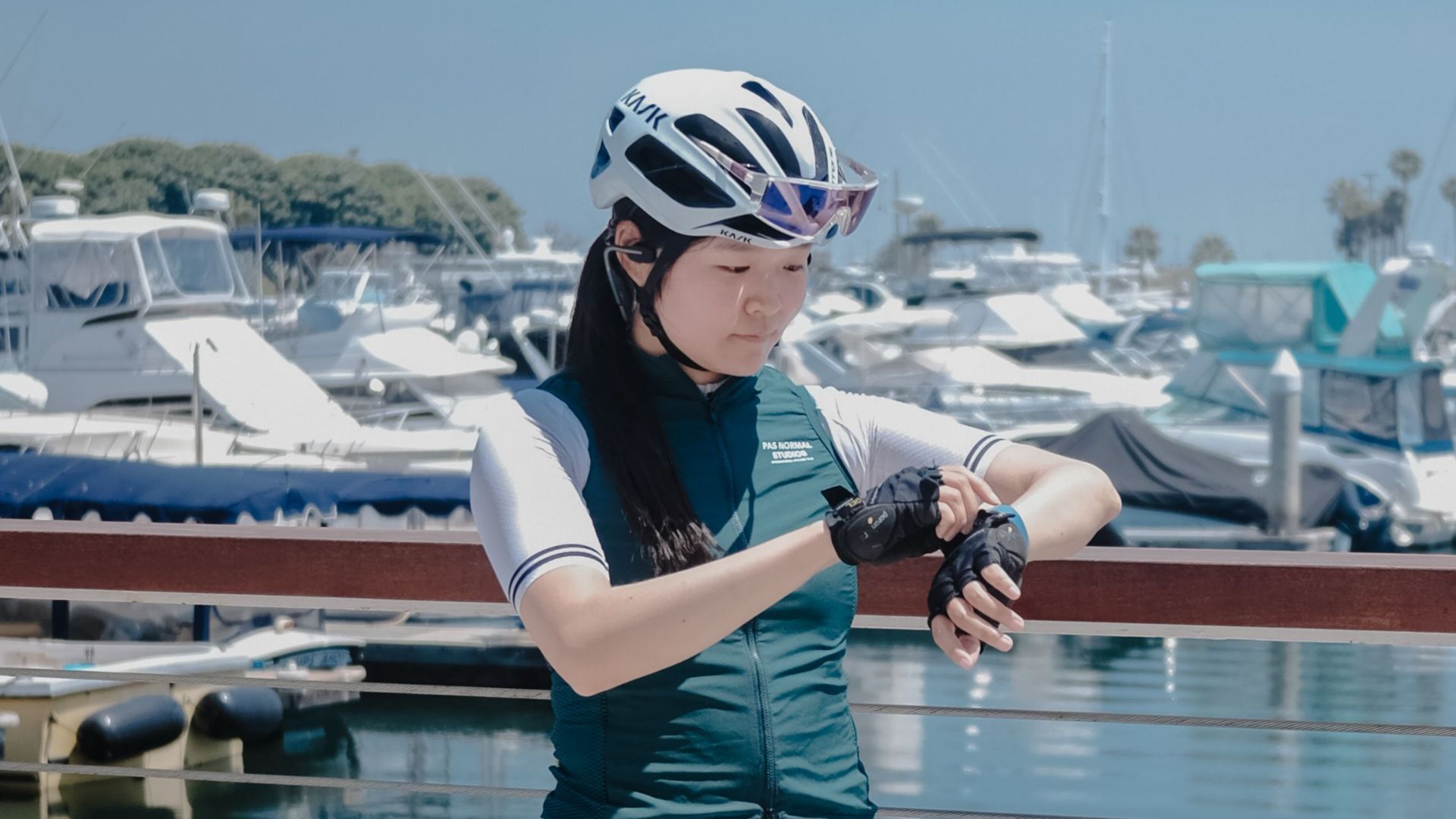 Athletic woman in cycling gear checking watch at marina harbor on a sunny day.