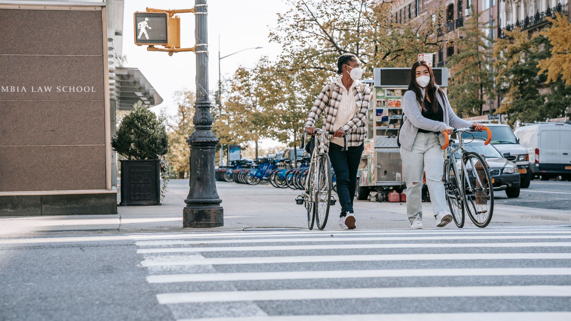 Unrecognizable young multiethnic female friends in masks with bikes in casual apparel strolling on crosswalk and talking in town