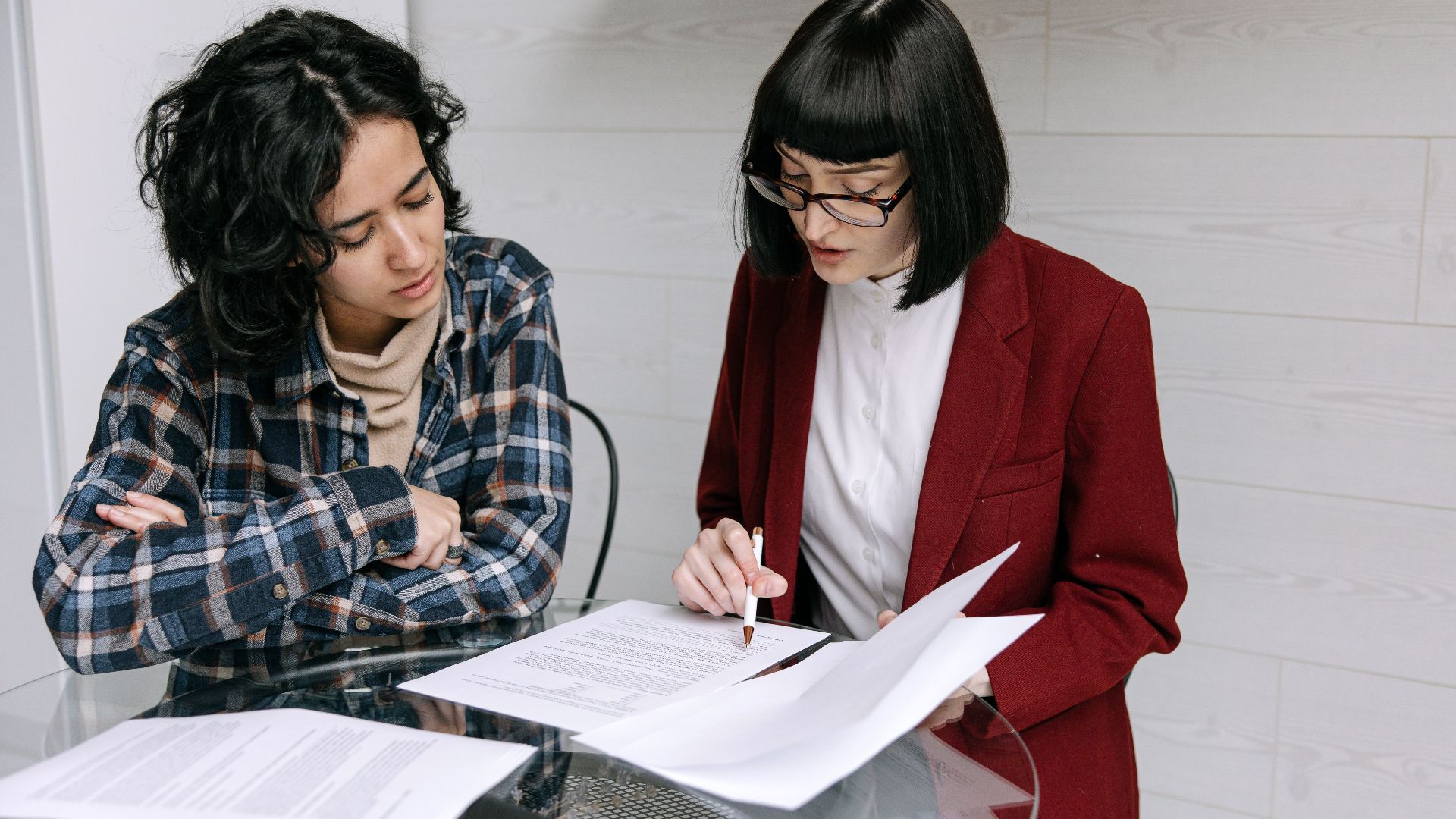 Two women collaborating at a glass table, reviewing business documents in a modern office setting.