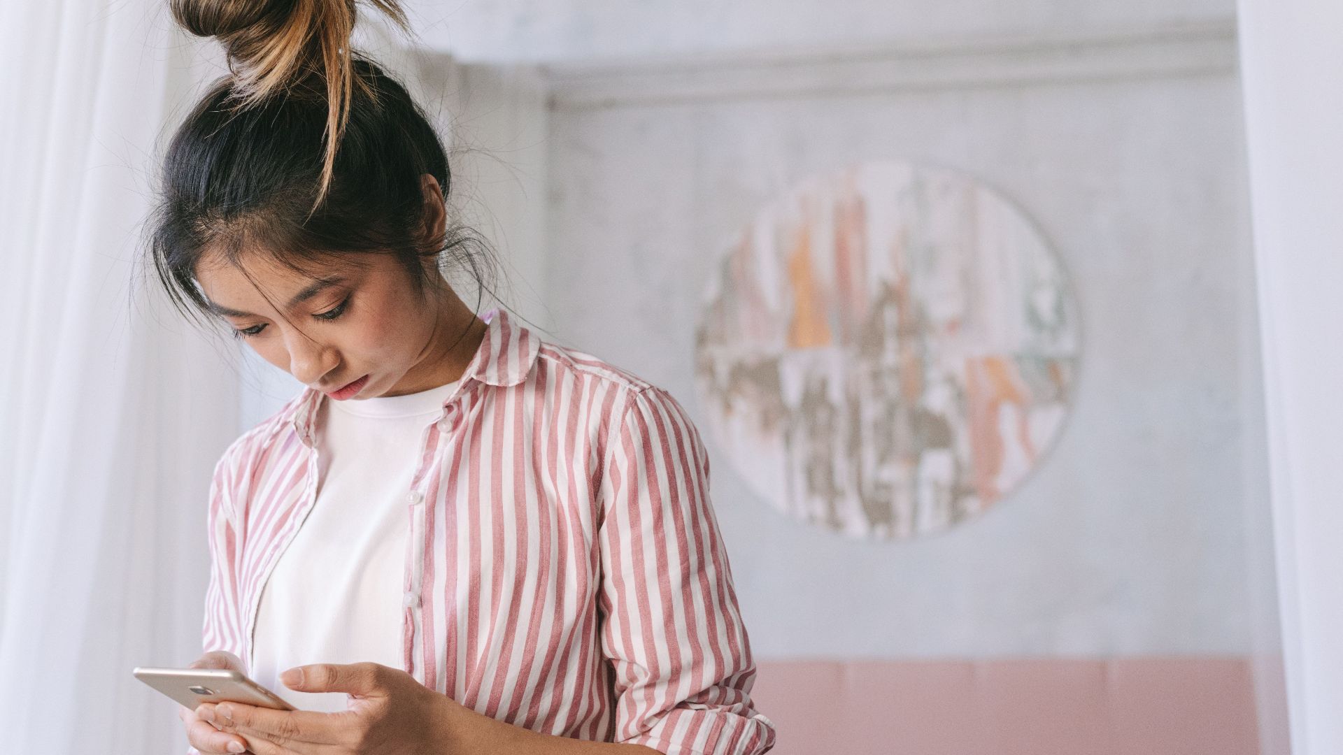 A young woman in a striped shirt using her smartphone indoors with soft decor and a relaxed atmosphere.