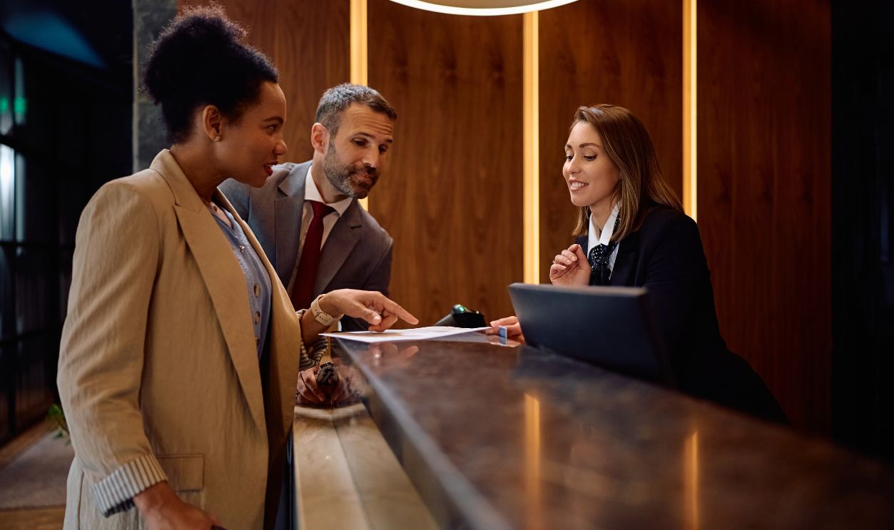 Shutterstock-2613366849, Happy hotel manager assisting guest with filling out paperwork at reception desk.