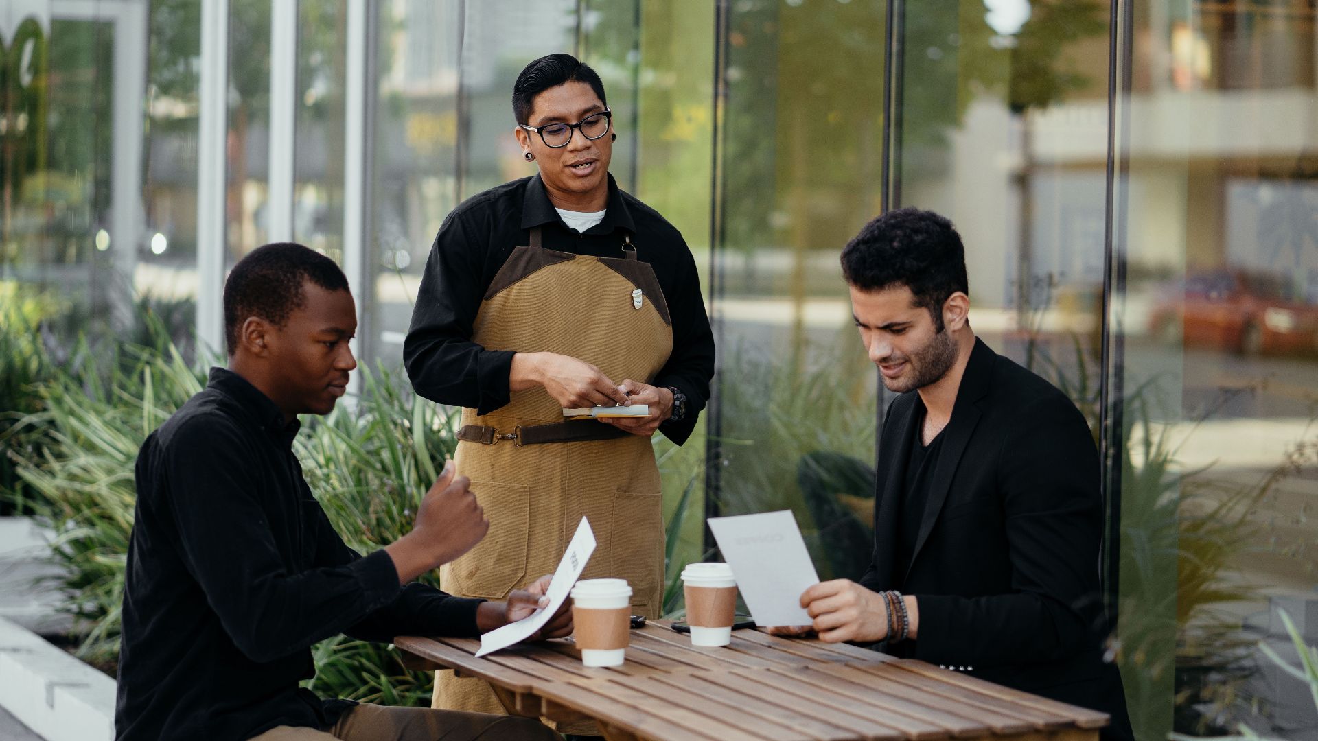 Two customers engaging with a waiter at an outdoor café table, reviewing the menu.
