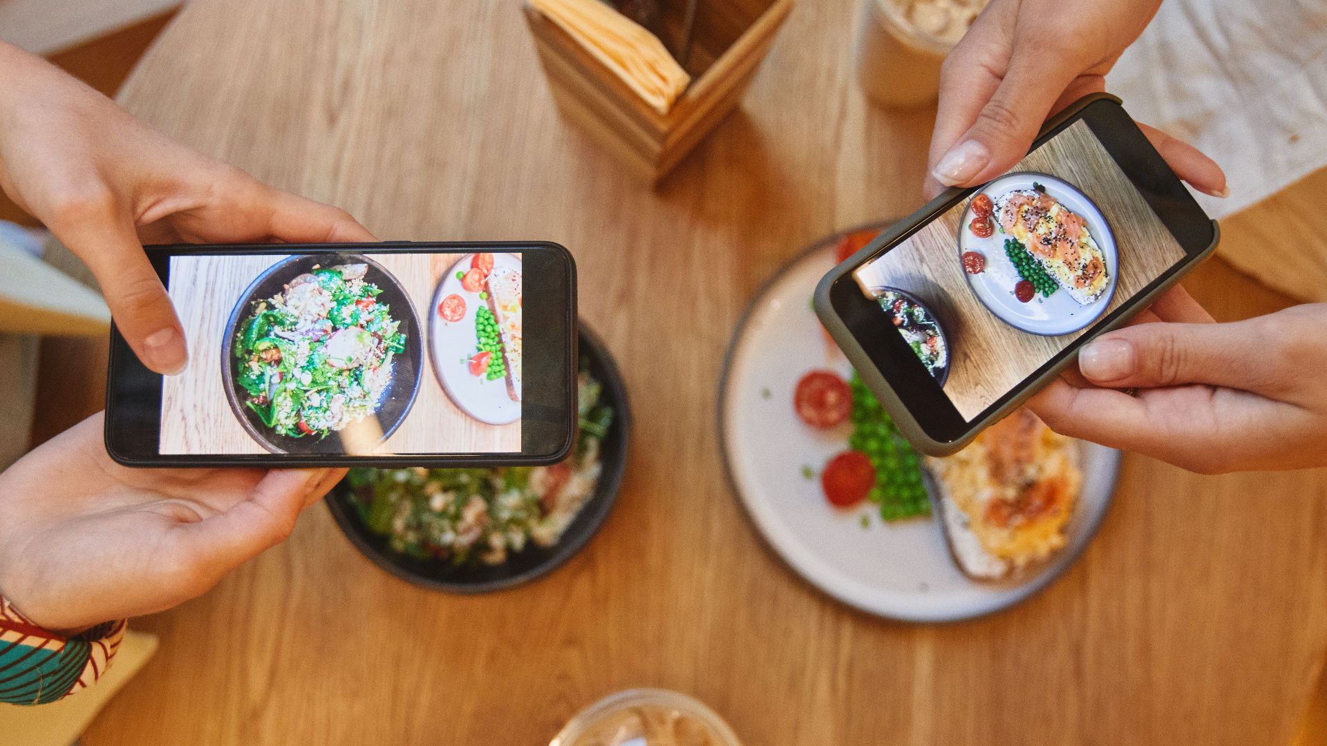 Two people taking photos of brunch dishes with smartphones in a cozy cafe setting.