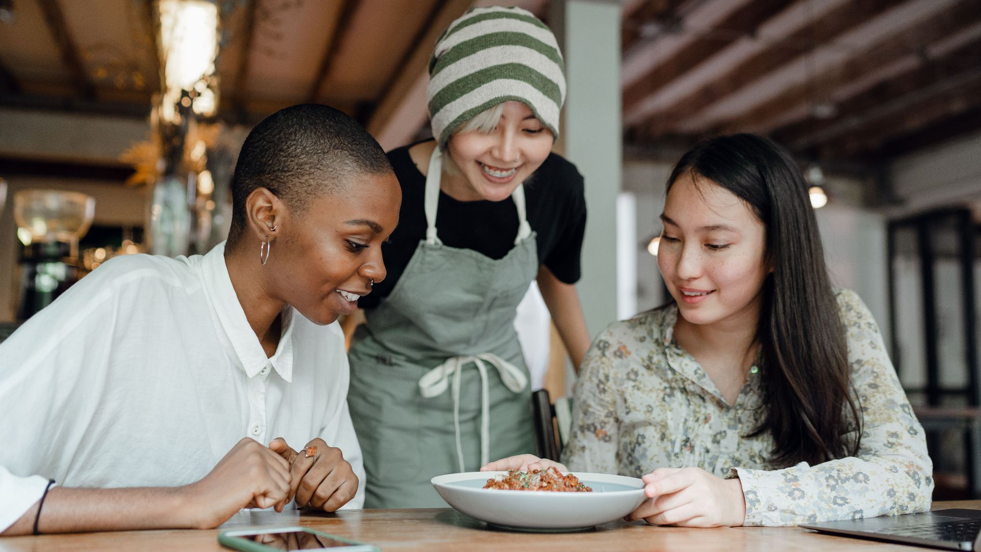 Smiling positive multiracial females in casual clothes sitting at table in modern cafe near cheerful waitress and looking at delicious dish