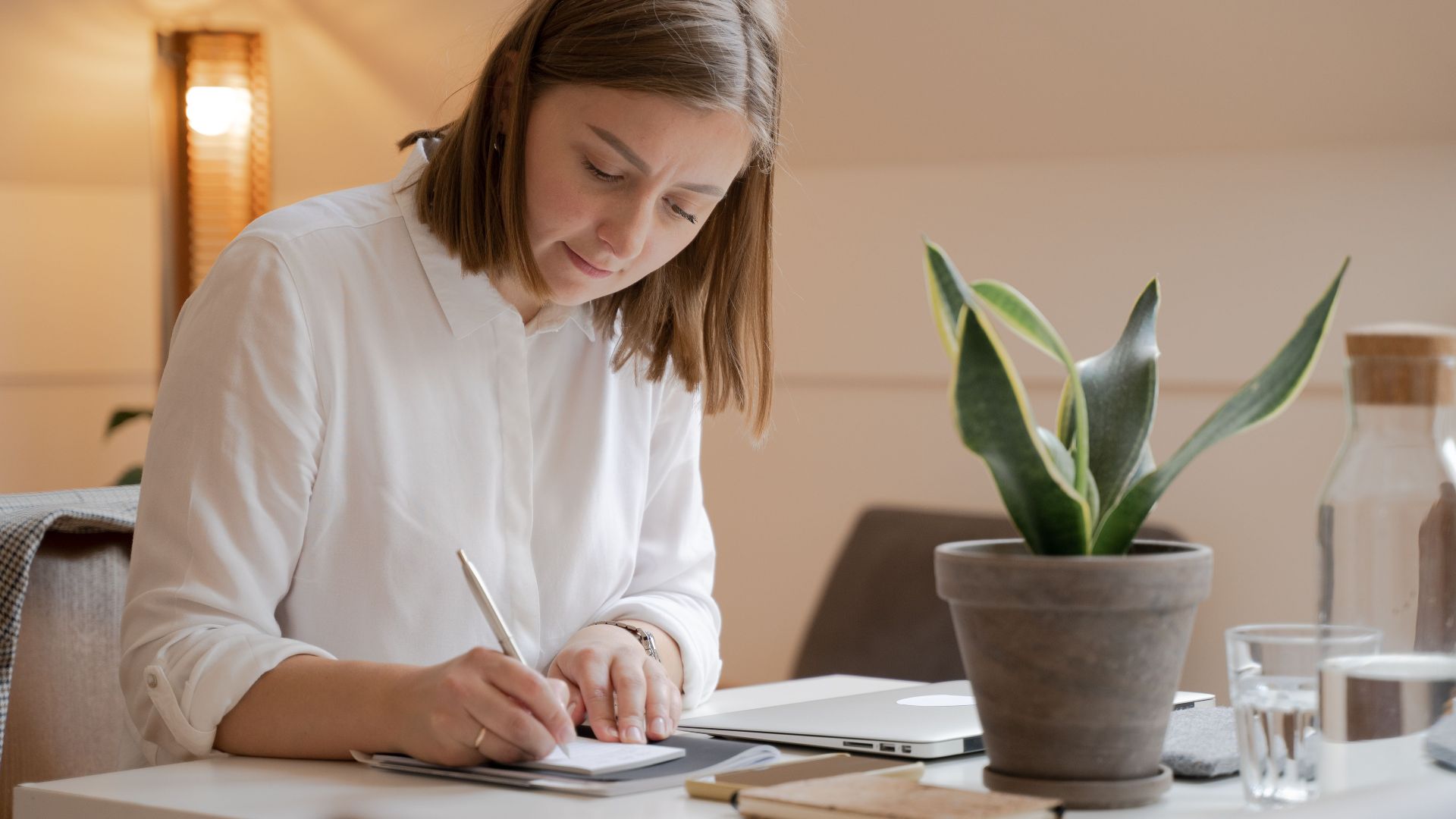 Woman in white shirt writing at a desk with a potted plant, creating a calm work environment.