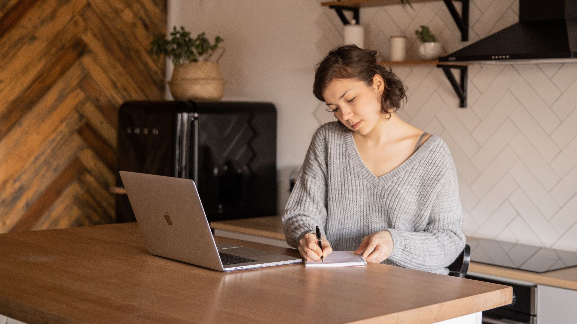 Content young businesswoman in gray sweater sitting at table with netbook in modern kitchen and taking notes while working on remote project in home workplace