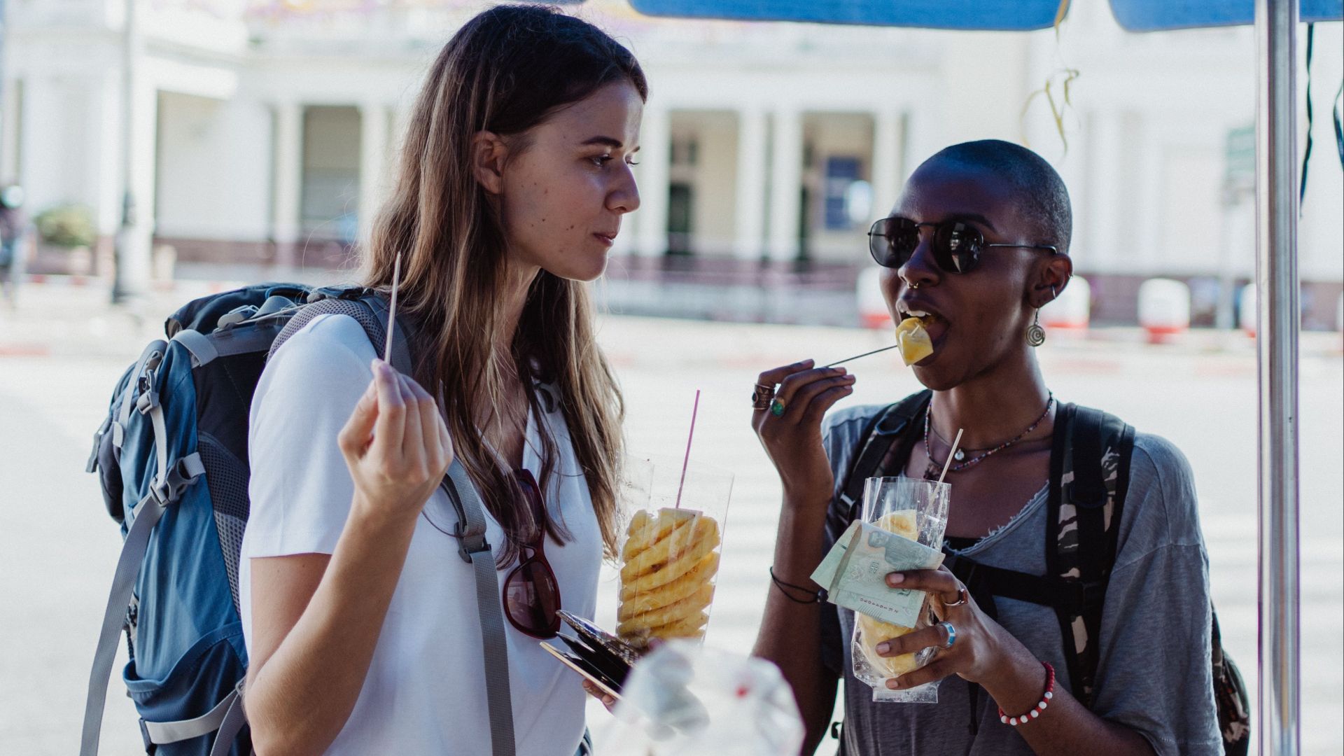 Two travelers eating fresh pineapple from street vendors in a lively urban setting, enjoying a summery day.