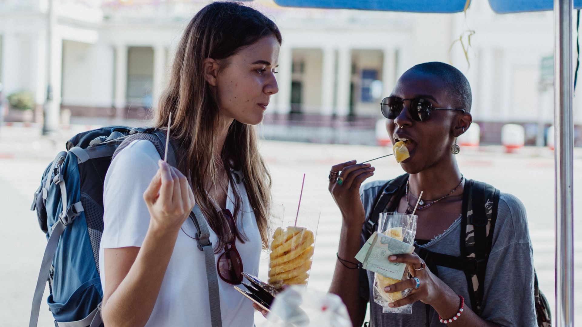 Two travelers eating fresh pineapple from street vendors in a lively urban setting, enjoying a summery day.