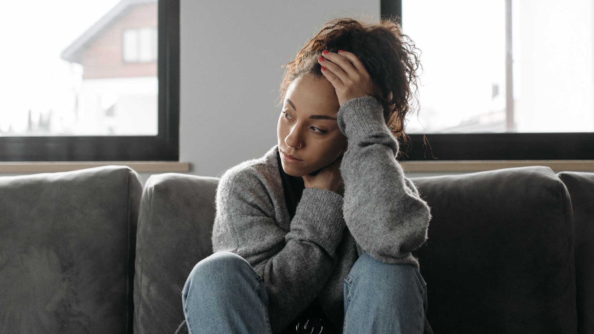 Thoughtful woman in cozy sweater sitting on sofa indoors by the window.