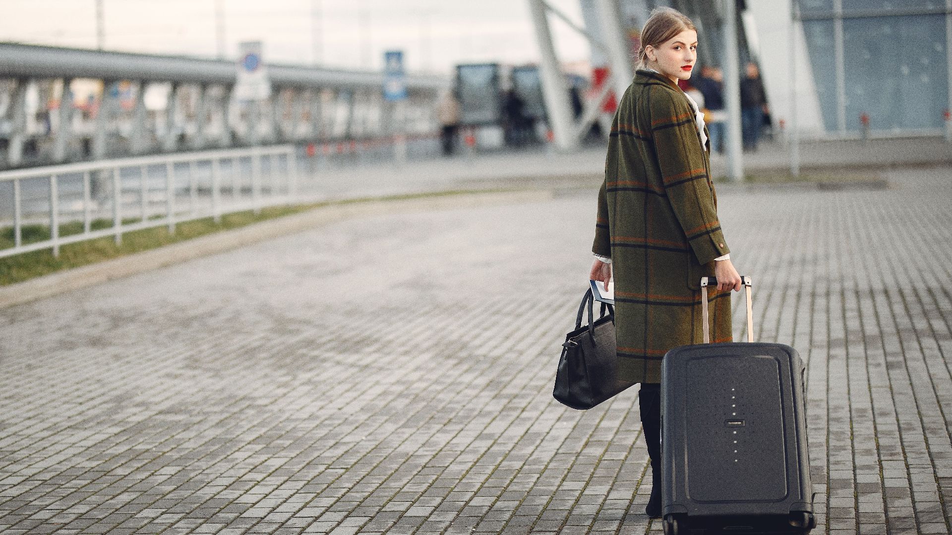 Full length of stylish female traveler in warm coat walking with suitcase and bag looking at camera after arrival against airport terminal