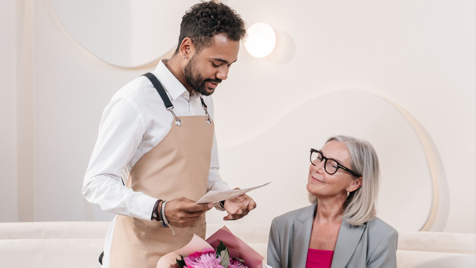 A waiter delivers a floral bouquet to a woman at a table in a stylish café setting.