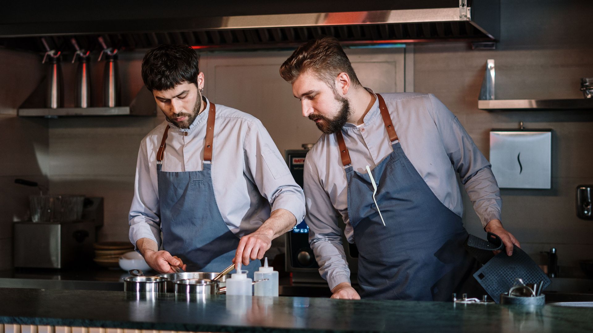 Two male chefs in aprons cook attentively in a contemporary open kitchen setting.