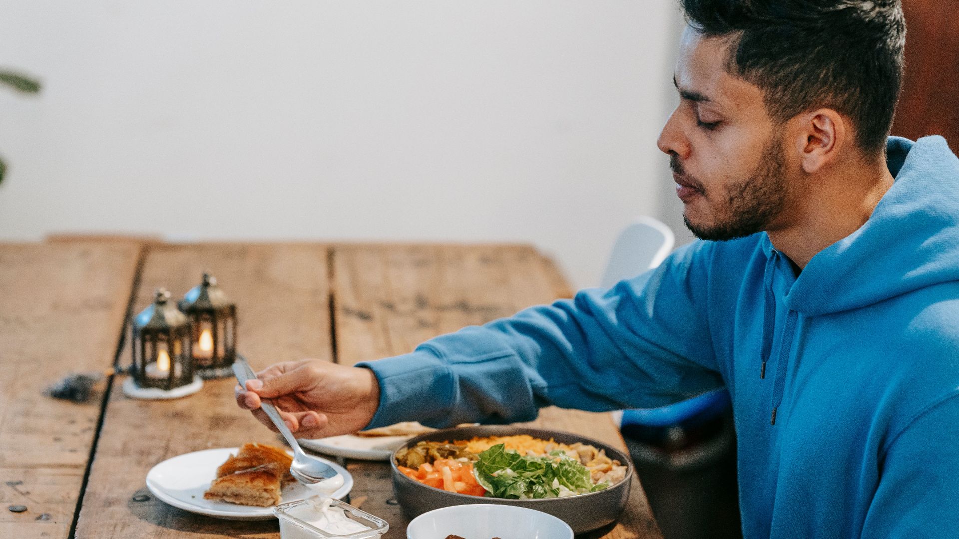 Side view of ethnic male in blue hoodie eating dessert and tasty meal at wooden table