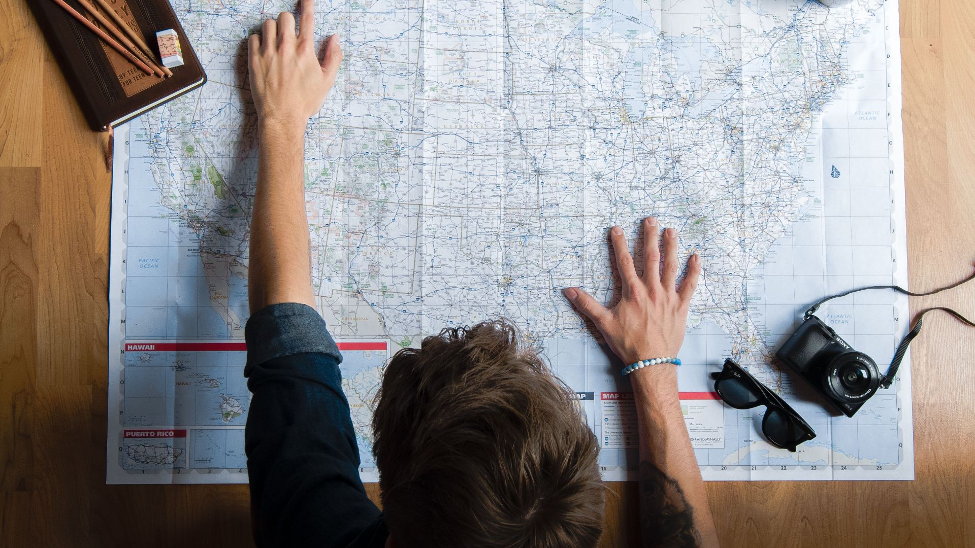 Top view of anonymous young male in casual clothes sitting on wooden floor with photo camera and coffee cup and pointing on paper map while planning trip route