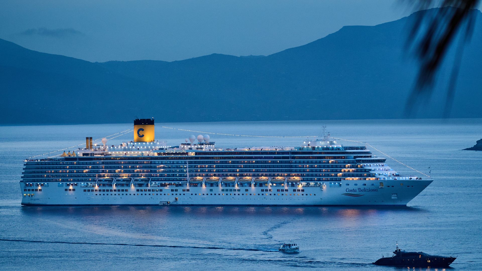 A beautifully illuminated cruise ship sails across the calm sea at dusk, framed by mountains.
