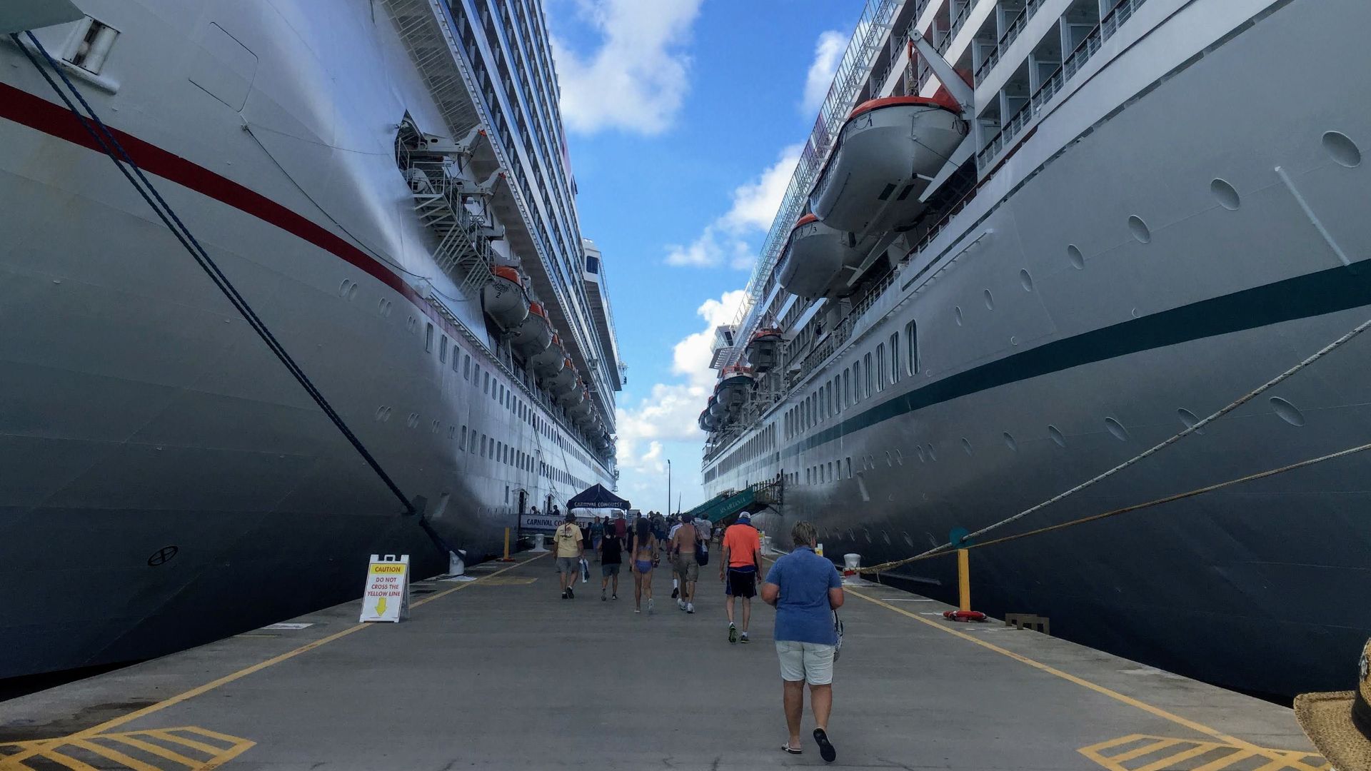 Tourists walking between massive cruise ships in a sunny harbor, enjoying a vacation experience.