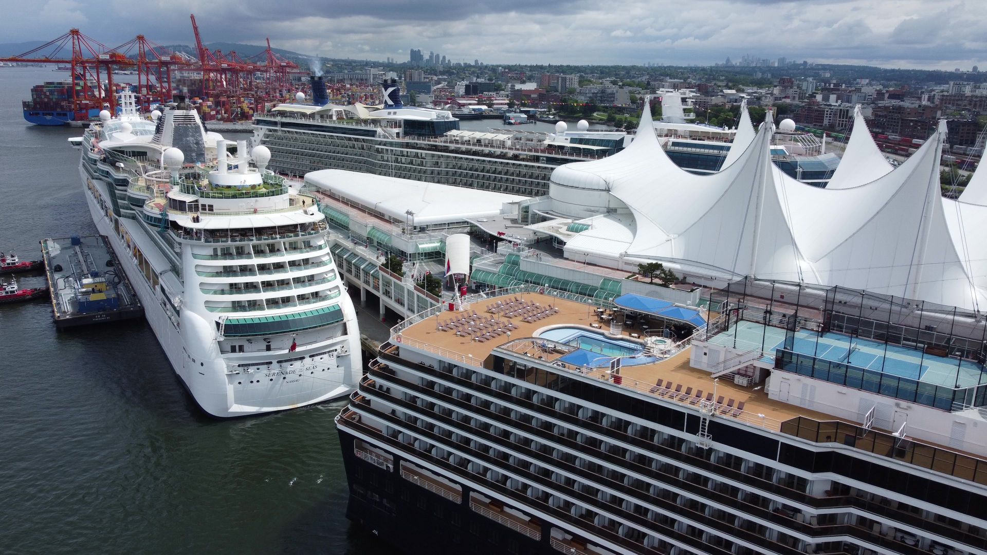 Aerial shot of two large cruise ships docked at a bustling port terminal under cloudy skies.