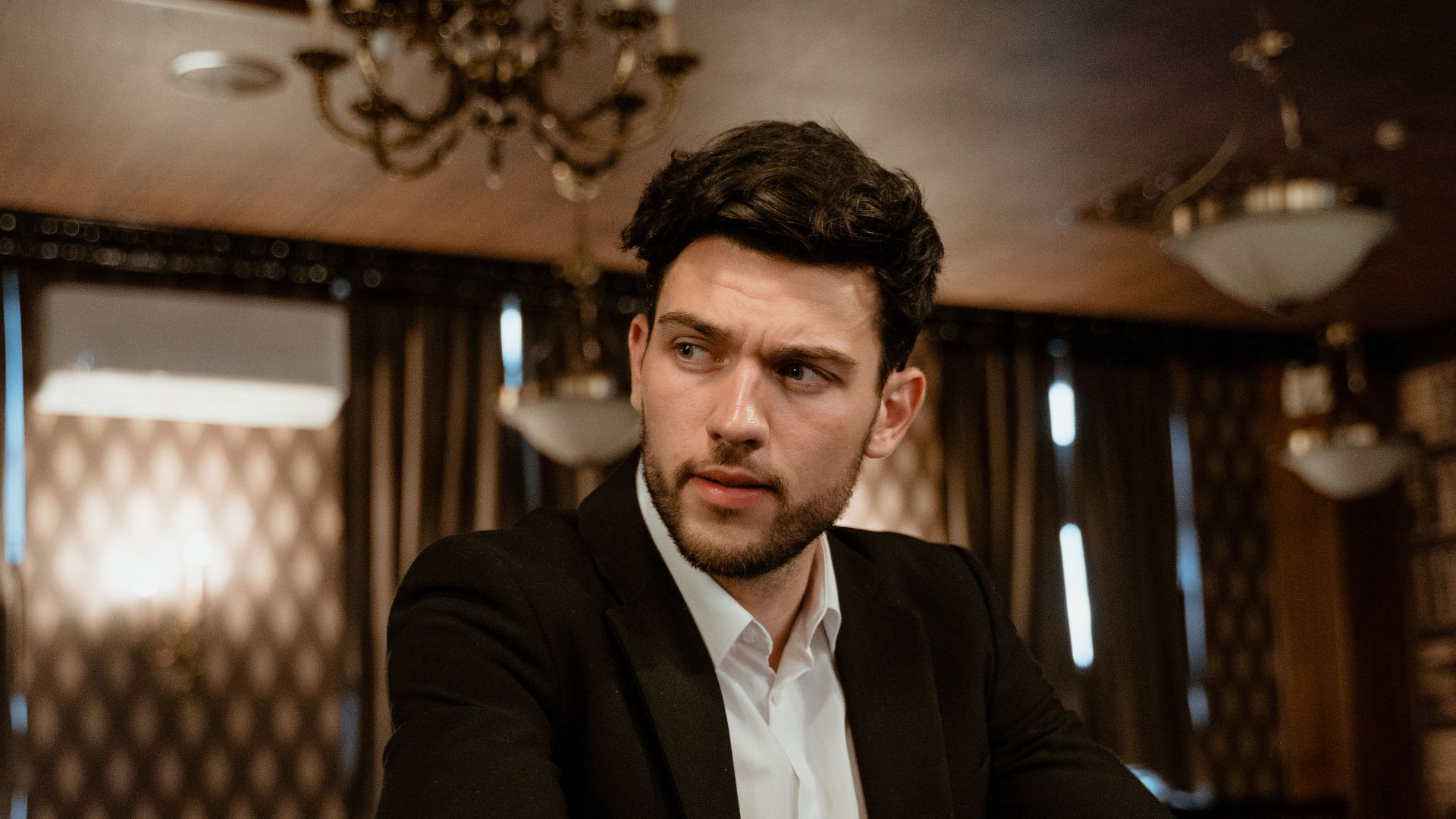 A man in a black suit with a drink, sitting at a bar in a sophisticated setting.