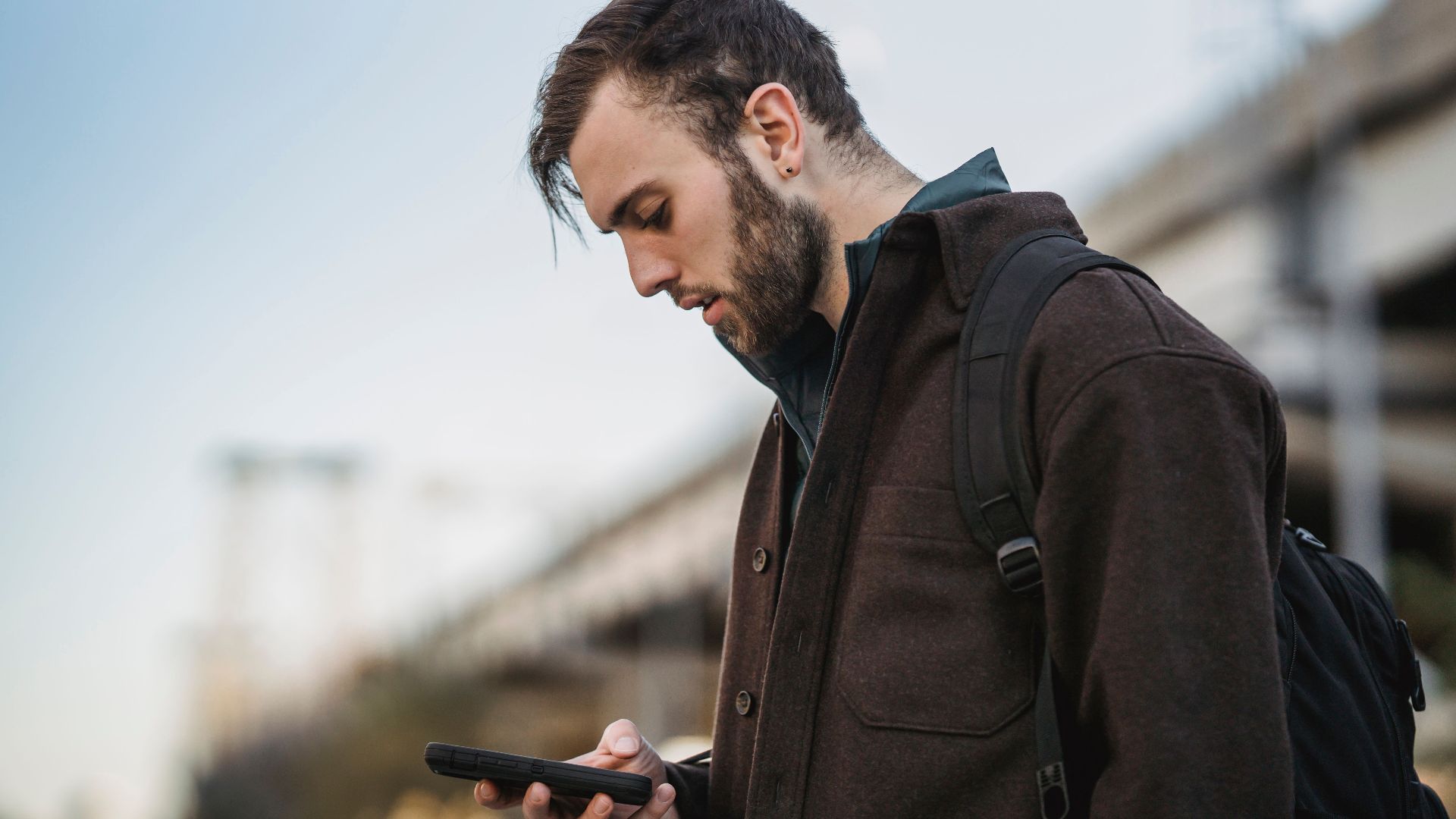 Side view of crop young unshaven male watching cellphone under light sky in town