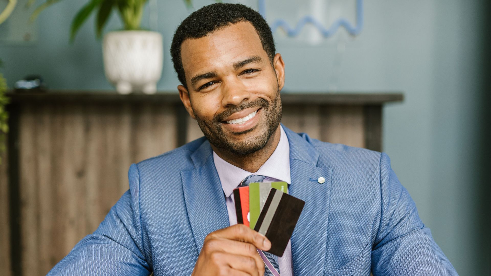 A confident businessman in a blue suit holds and displays credit cards while smiling at a desk.