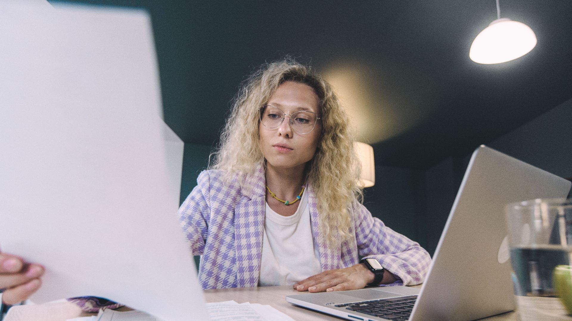 Woman in an office environment reviewing documents with focus, surrounded by technology.