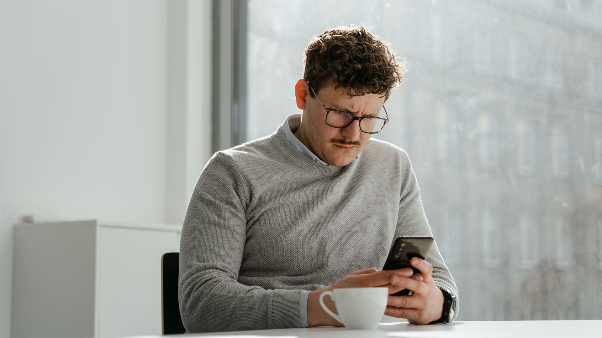 Adult man wearing glasses using smartphone in sunny office space, holding a mug.