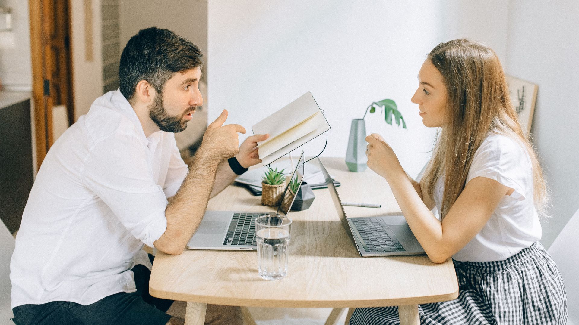 A man and woman working from home on laptops, engaged in discussion at a wooden table.