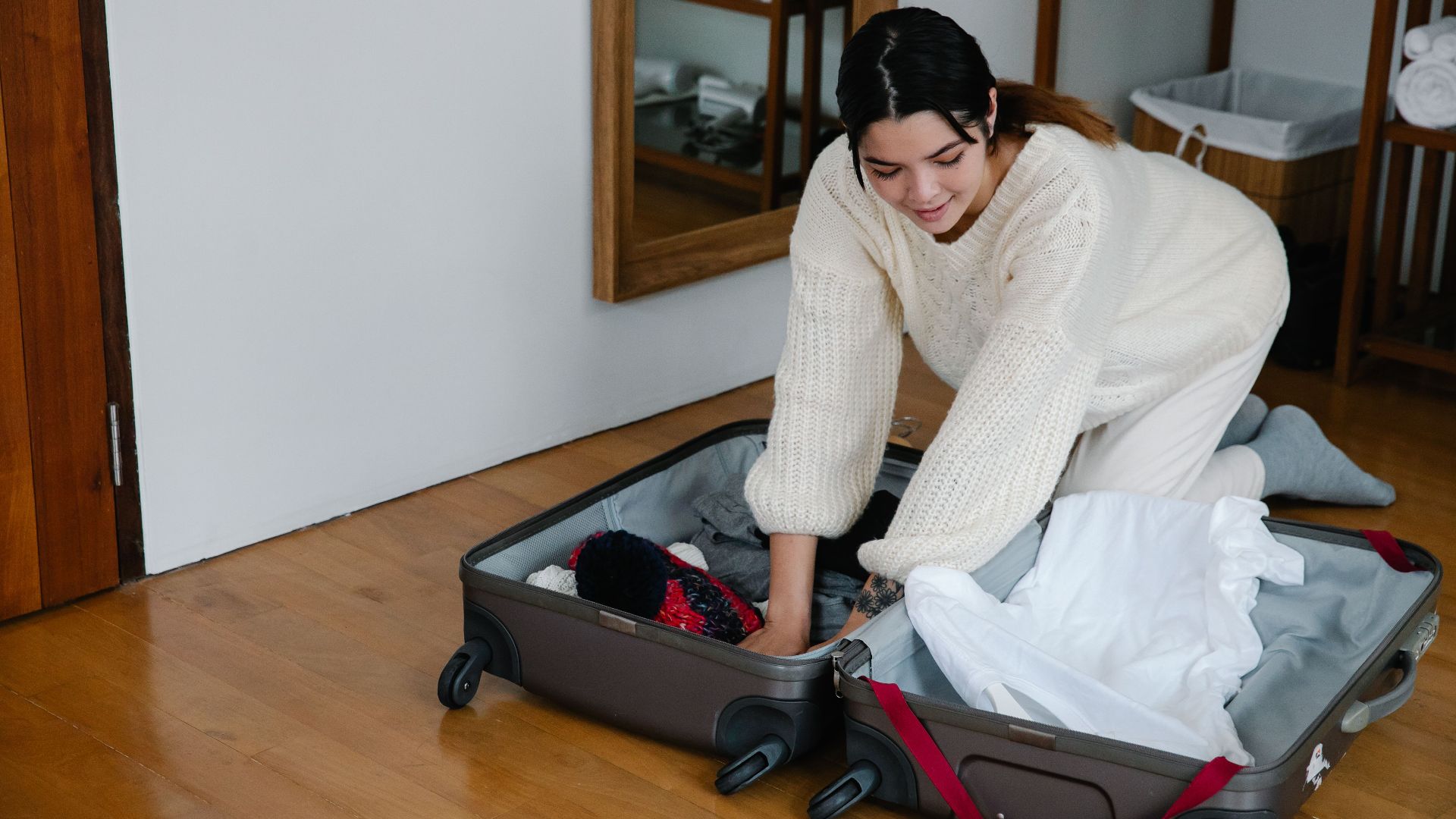 A woman kneels on the floor packing clothes into a suitcase in a cozy bedroom setting.