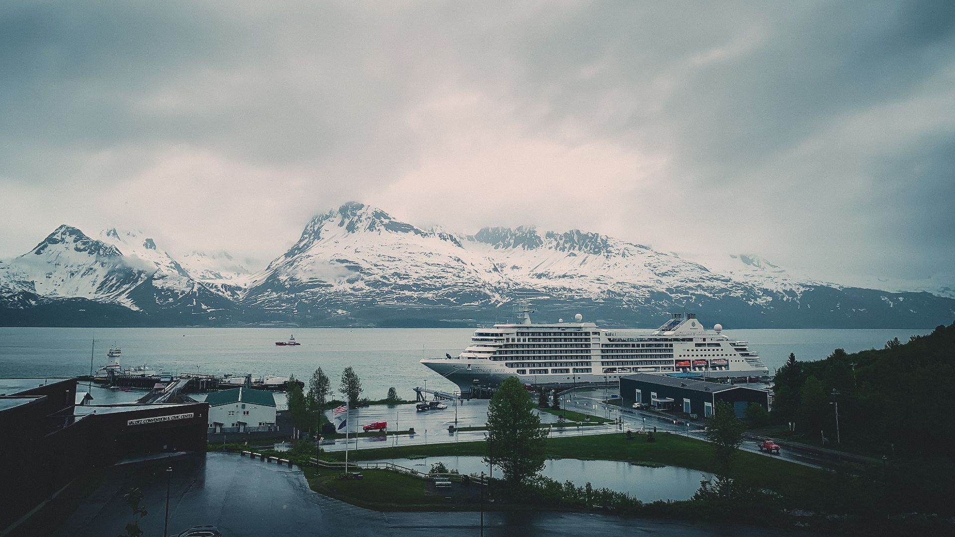 A cruise ship docked with snowy mountains in the background, under a cloudy sky.