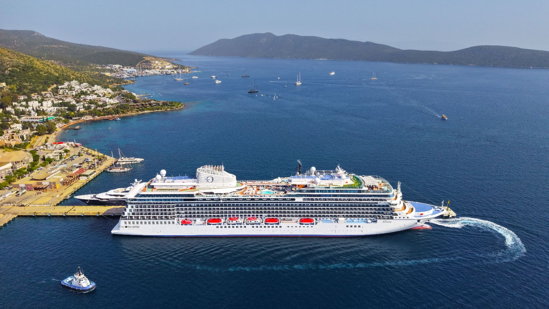 Aerial shot of a large cruise ship at a seaside port with beautiful coastal scenery in the background.