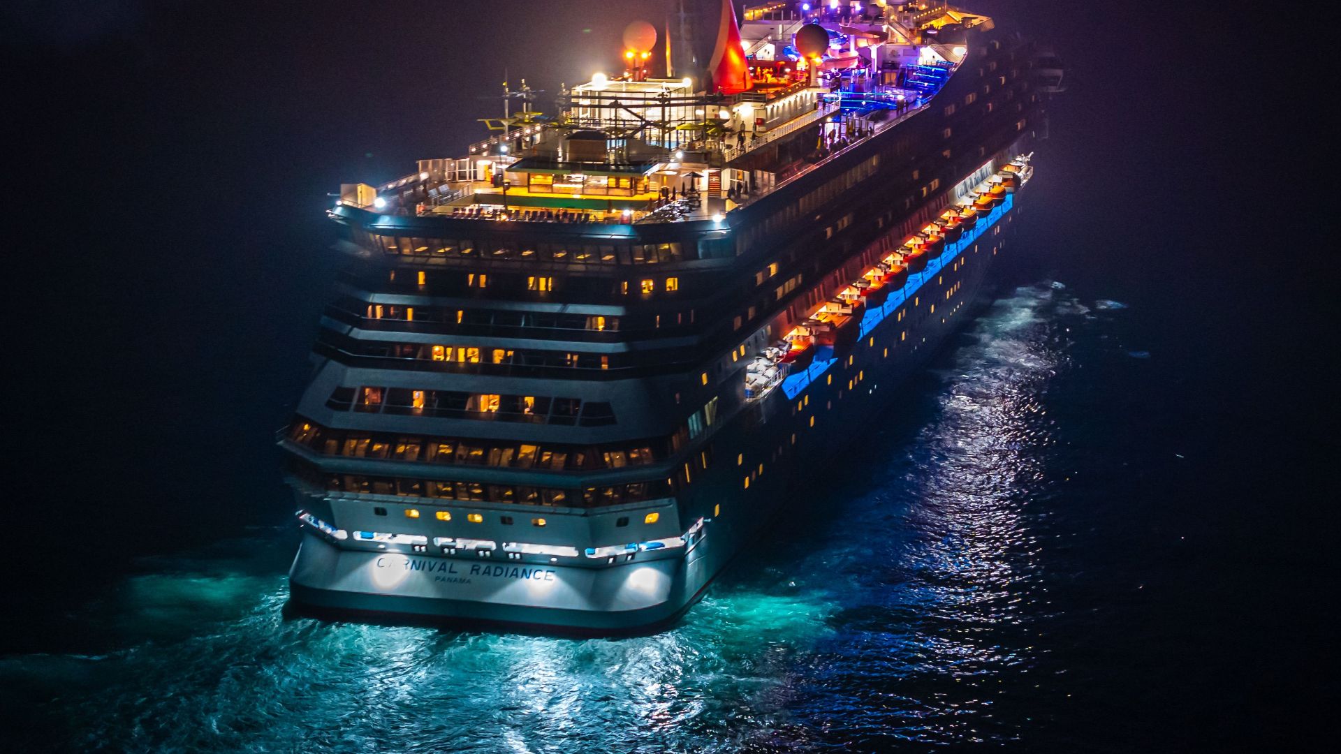 A vividly lit cruise ship sails through dark waters at night, creating a striking contrast.