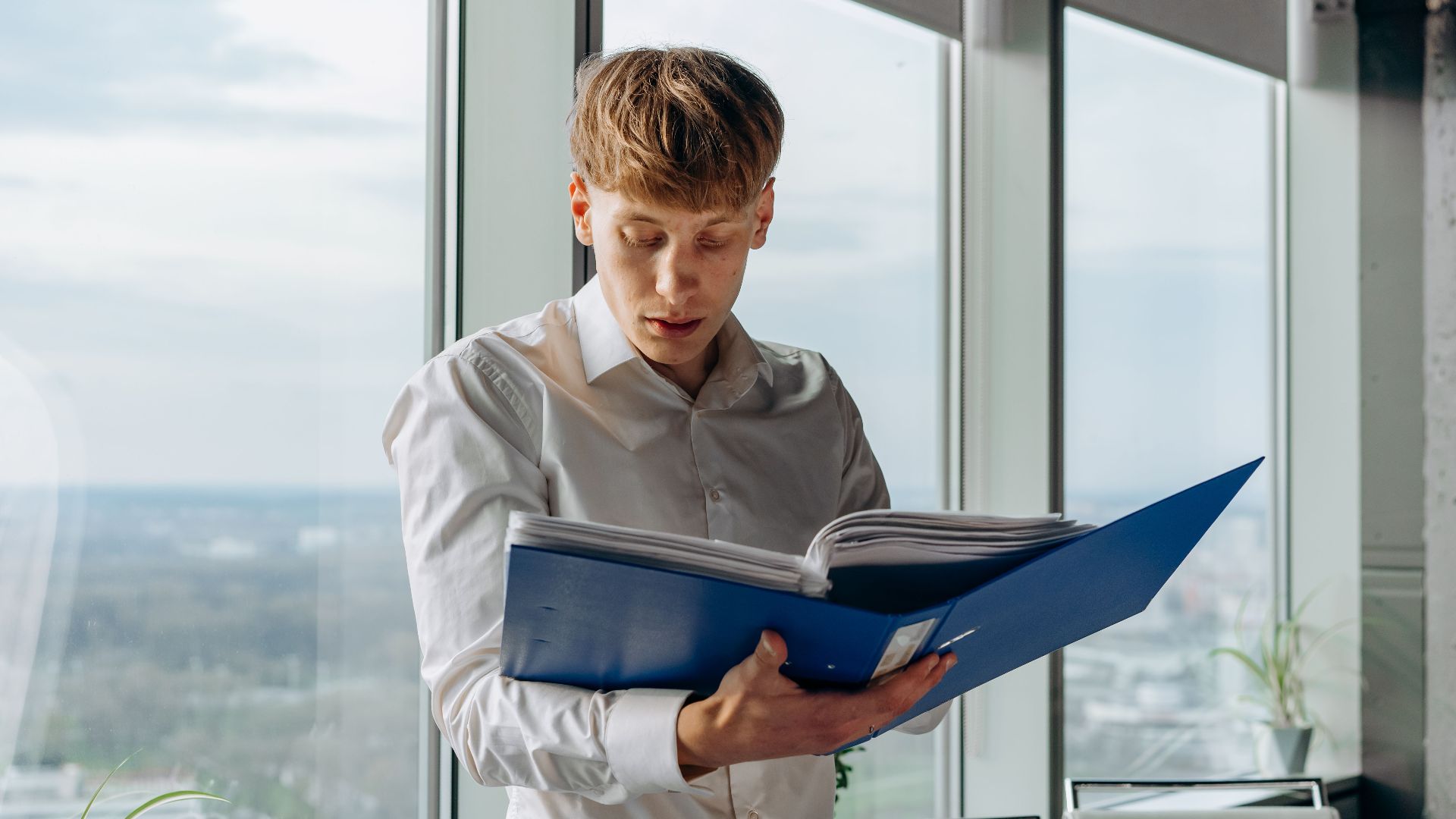 Businessman reading documents in an office with a city view, holding a blue binder.
