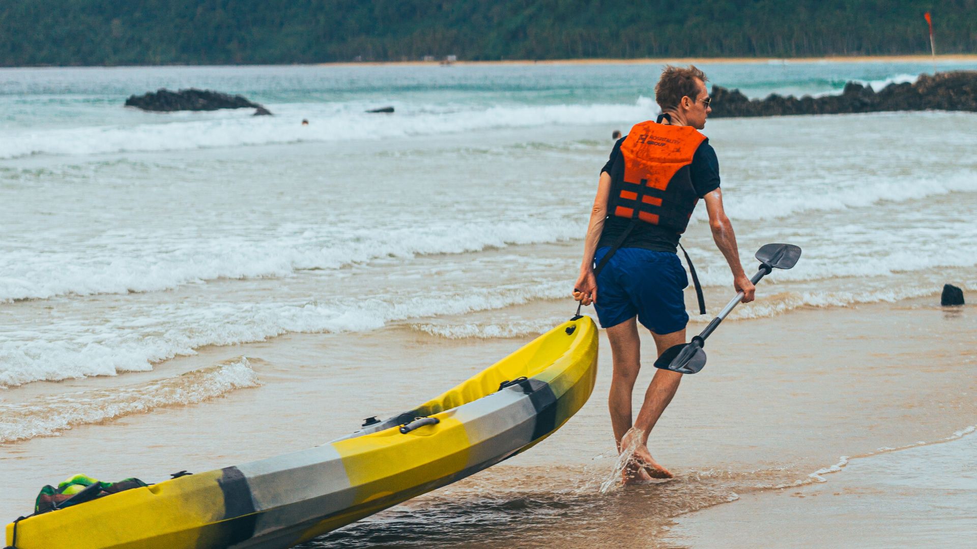 A man in a life jacket prepares to kayak on a tropical beach with lush hills in the background.