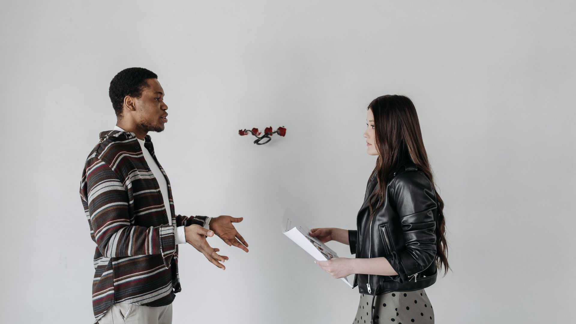 A man and woman in stylish outfits having a discussion in an interior setting with a white wall.