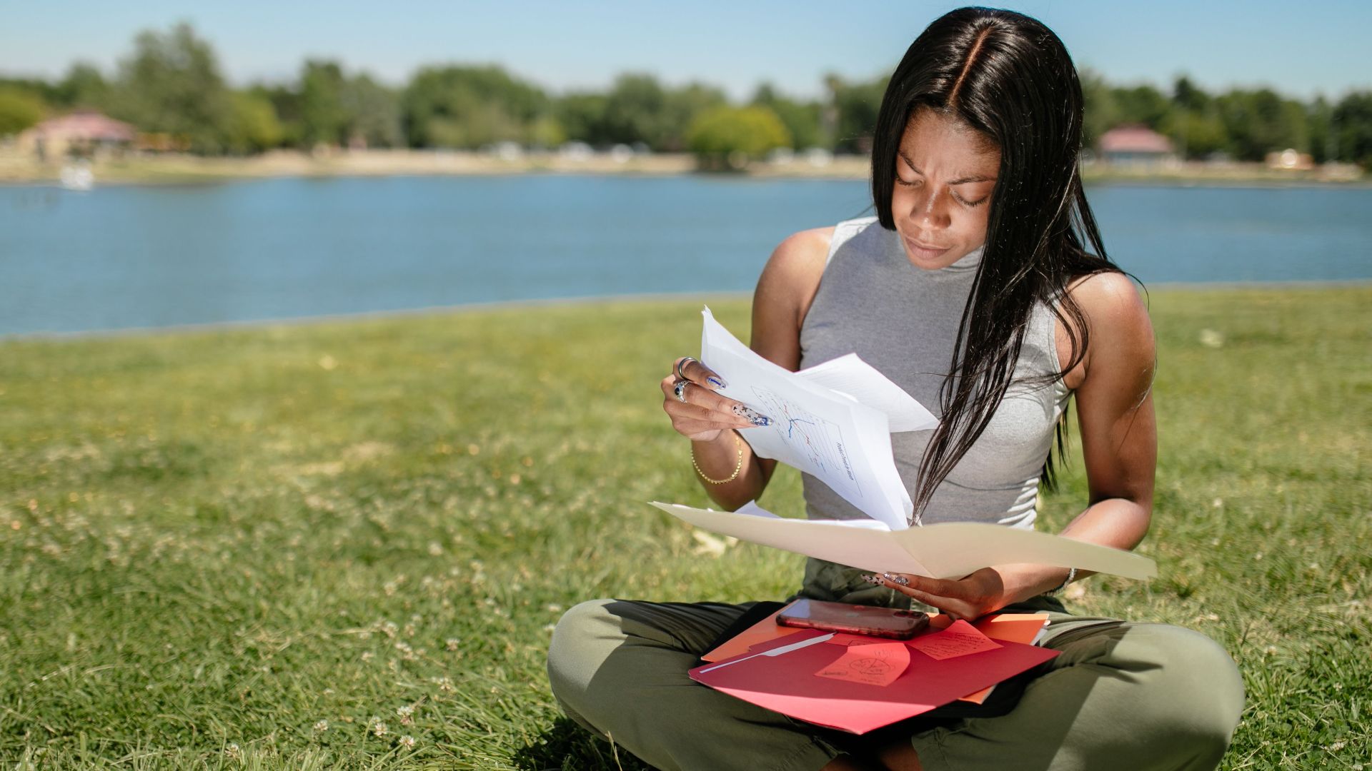 A woman sits on grass by a lake, examining papers on a sunny day.